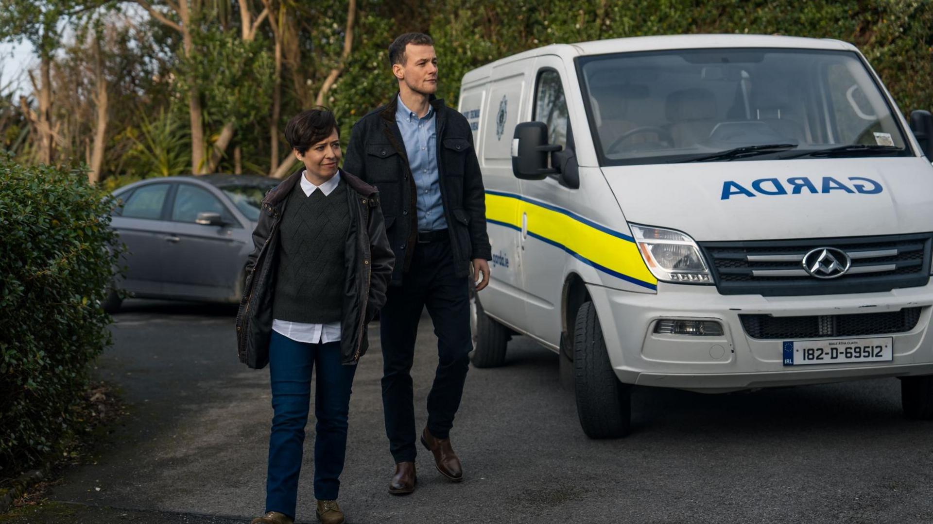 A woman - Caoimhe Farren - and a man - Alan Mahon - are walking on a driveway parked beside a white Garda van. The woman is wearing a short coat, a white collared shirt with a green jumper over the top. Beside her is a taller man wearing a blue shirt, a dark coat and trousers.