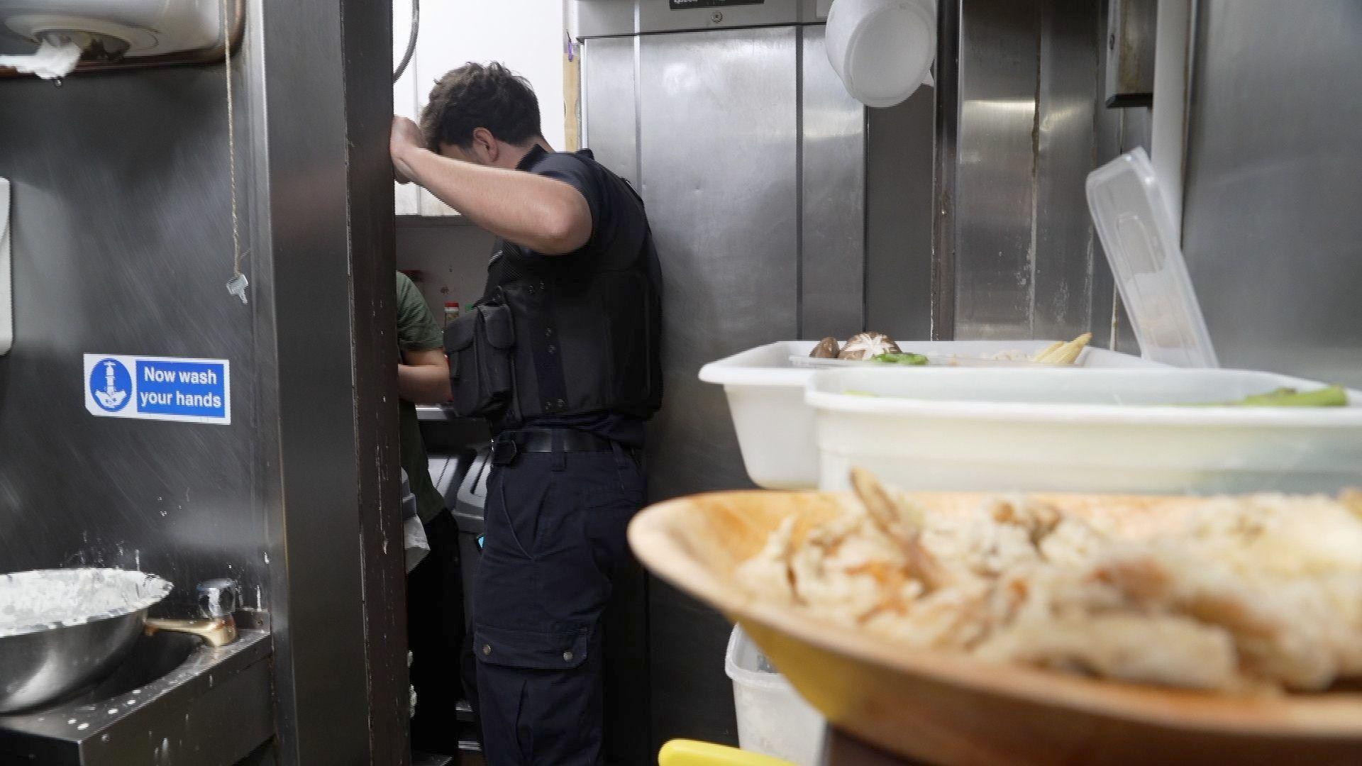 A kitchen where there are bowls of food in white plastic containers and a wooden bowl on the left side on a counter and then an immigration officer standing at the doorway of the kitchen leaning into another area where you can see the elbow and t-shirt of the person who is he speaking to. On the left, in the foreground is a metal sink with the sign 'now wash your hands' above it.