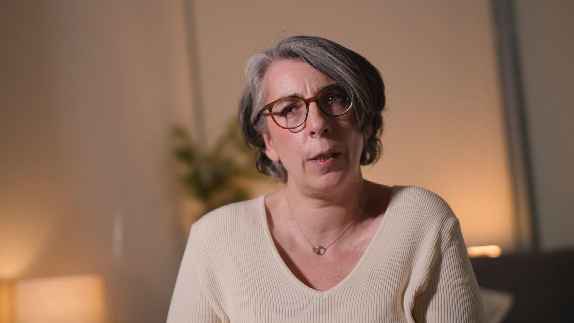 Bronagh Andrew has short, grey hair and glasses. She is wearing a beige ribbed jumper with a V-neck, showing a silver necklace. The background is softly lit and blurred.