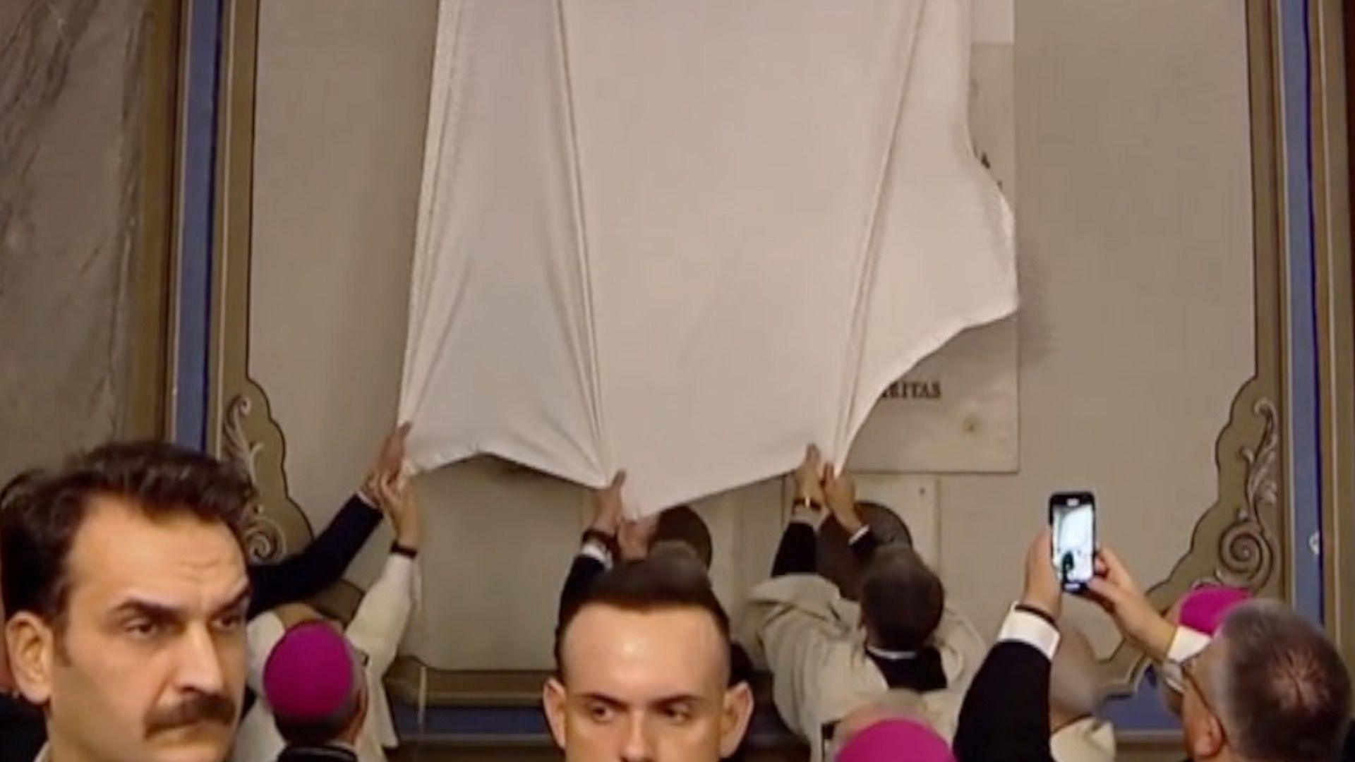The Pope and church staff tug at white fabric covering a plaque inside a church