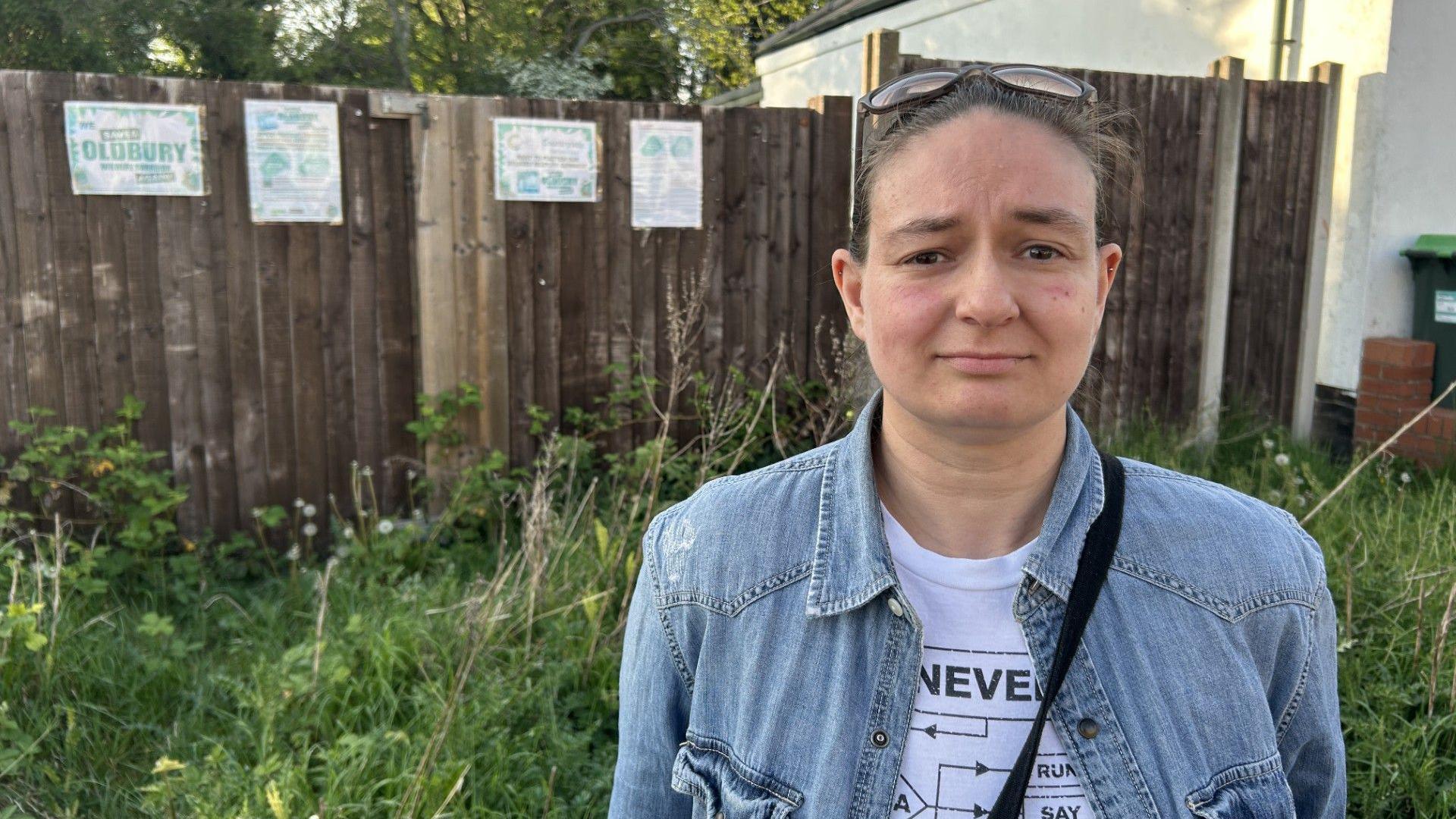 Tess Pegg standing next to signs opposing the Oldbury development. She has brown hair tied up and is is wearing a denim jacket and has sunglasses on her head. There is a fence behind her and some grass and weeds.
