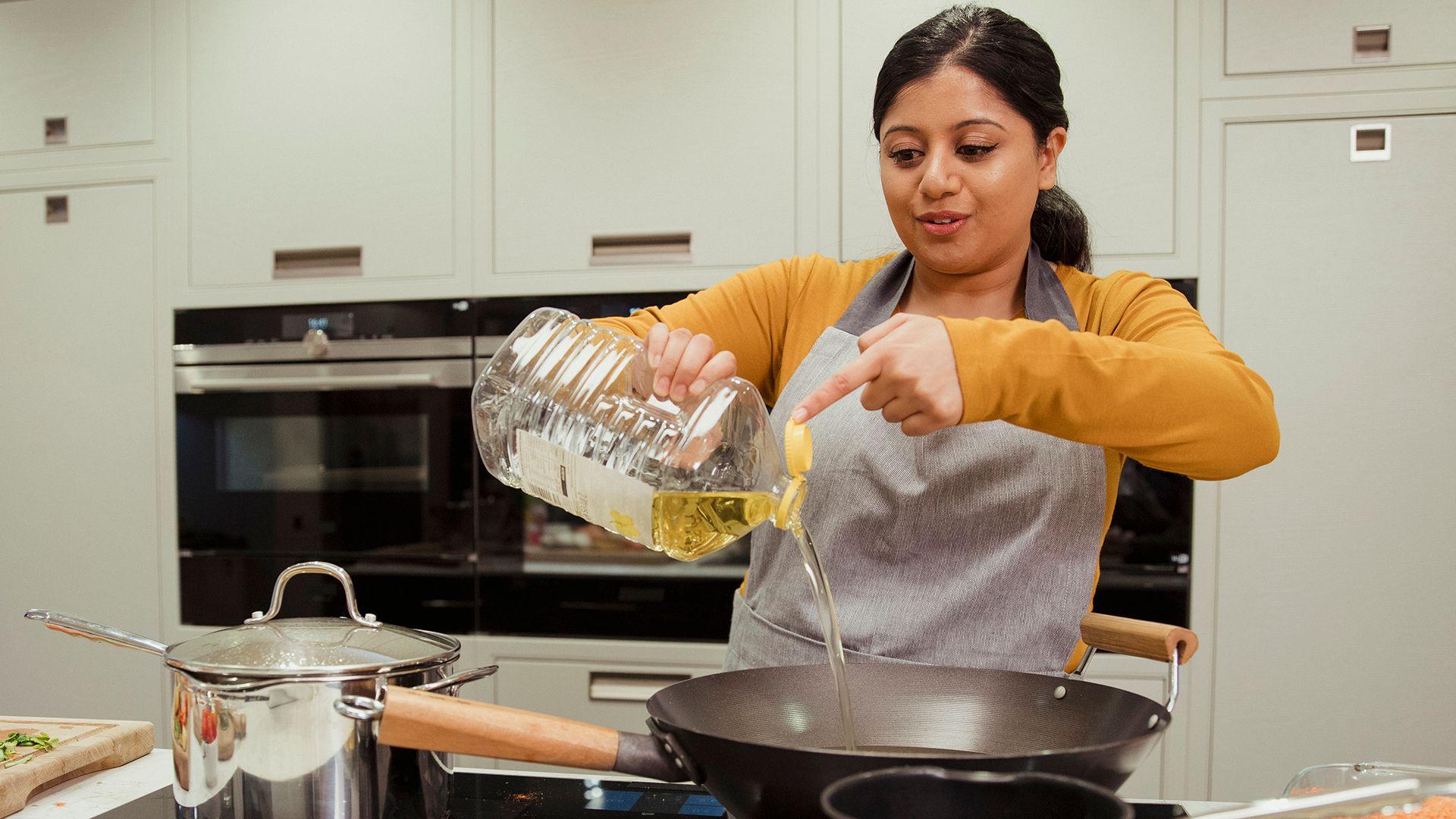 A stock photo shows a woman pouring a big bottle of sunflower oil into a deep wok pan on an electric hob with cooking ingredients all around her and her kitchen cabinets and oven in the background.