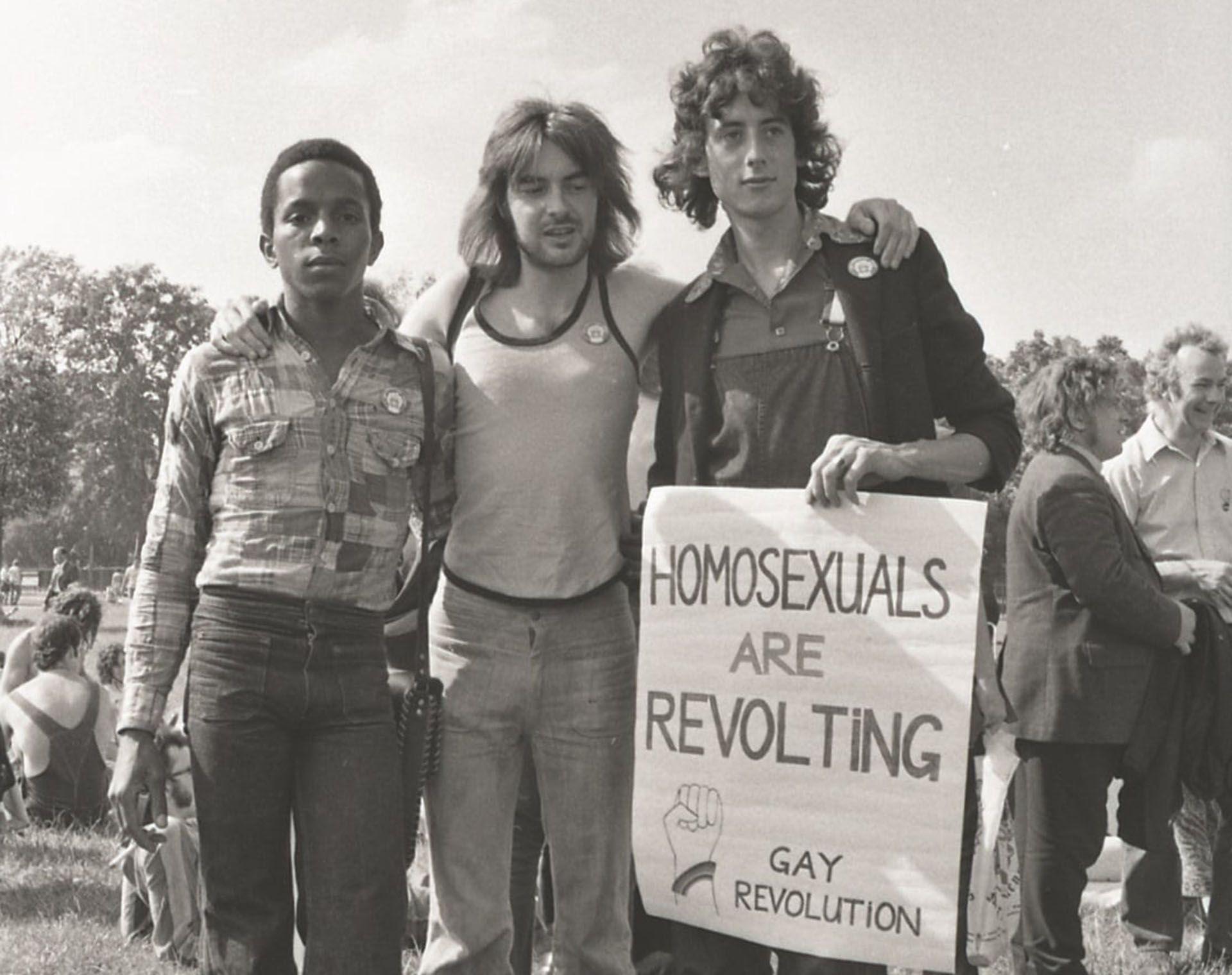 black and white picture of three men standing up in a park. one holds a sign reading homosexuals are revolting gay revolution