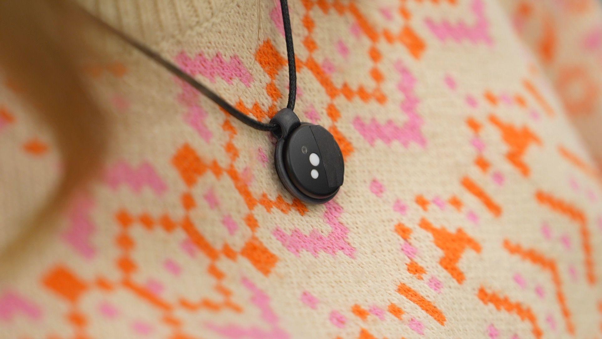 A pendant UV monitor is placed around someone's neck. It is on a black cord that looks a little like a thin round shoelace and is a plastic circular disk with three litte light monitors on the front of it. The information goes to an app.