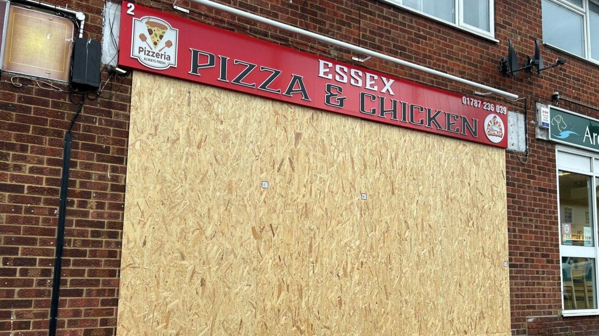 The front of Essex Pizza and Chicken shown completely boarded up with a large sheet of chipboard.