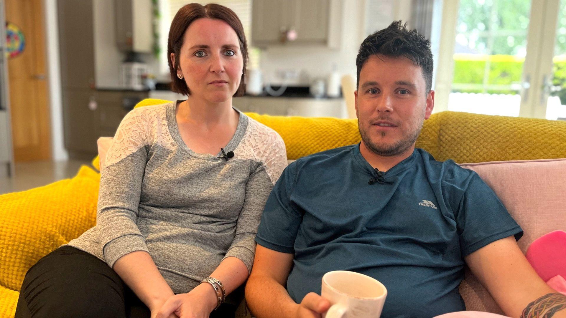 Man wearing dark blue shirt with black hair and stubble, sitting on a yellow sofa beside a woman with short brown hair wearing a grey top.