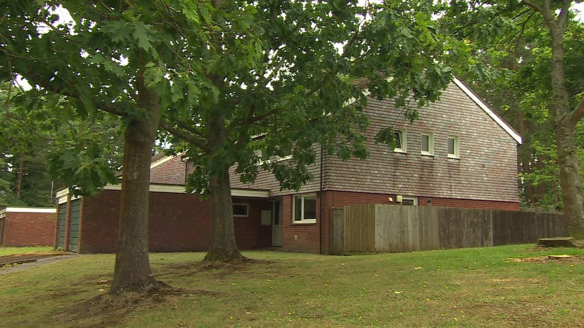 A two-storey house behind a green with two trees on it. The bottom storey is brick and the top storey is tiled shingles.