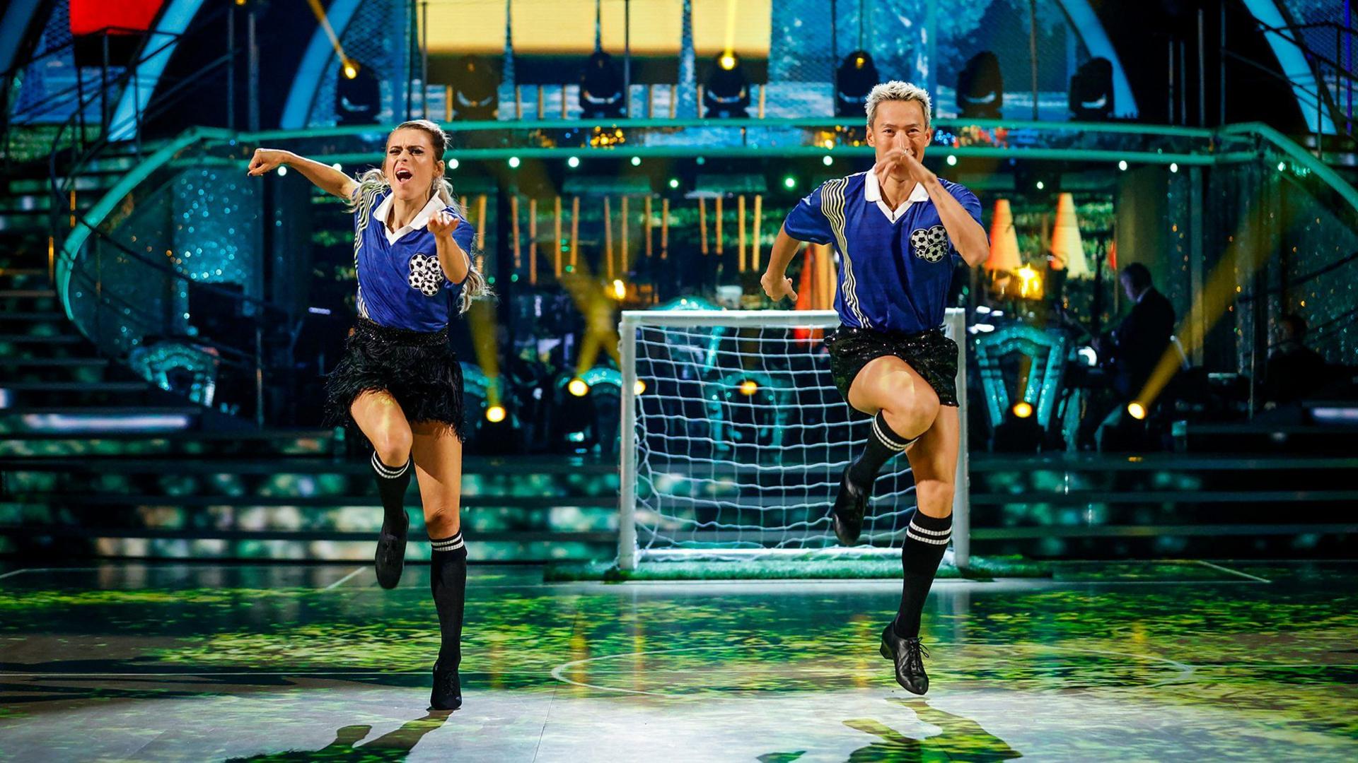 Karen and Carlos energetically dance a Jive, wearing matching football tops and shorts on a dancefloor designed like a pitch