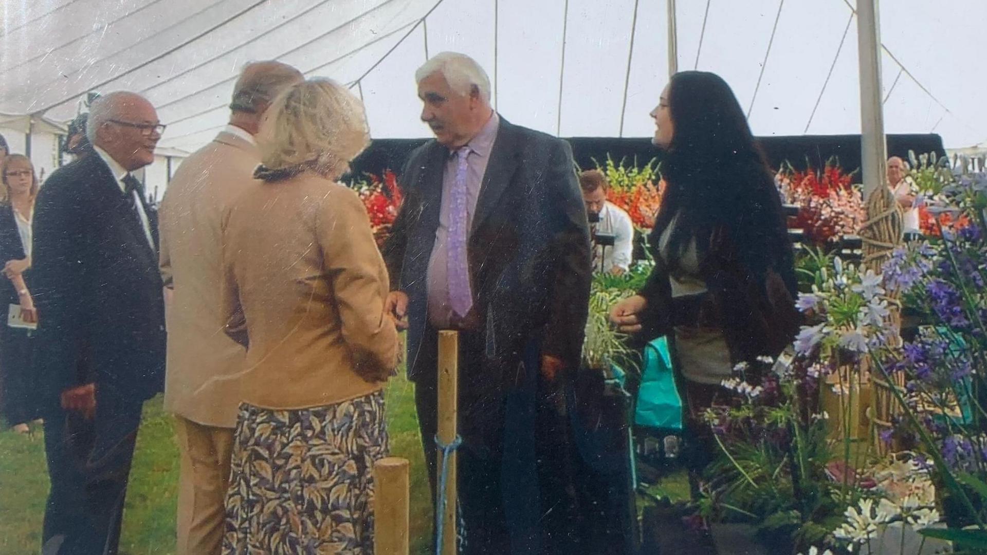 The picture shows a group of people in a marquee standing next to a flower display. King Charles and Queen Camilla can be seen from behind. They are chatting to people at the flower stall.