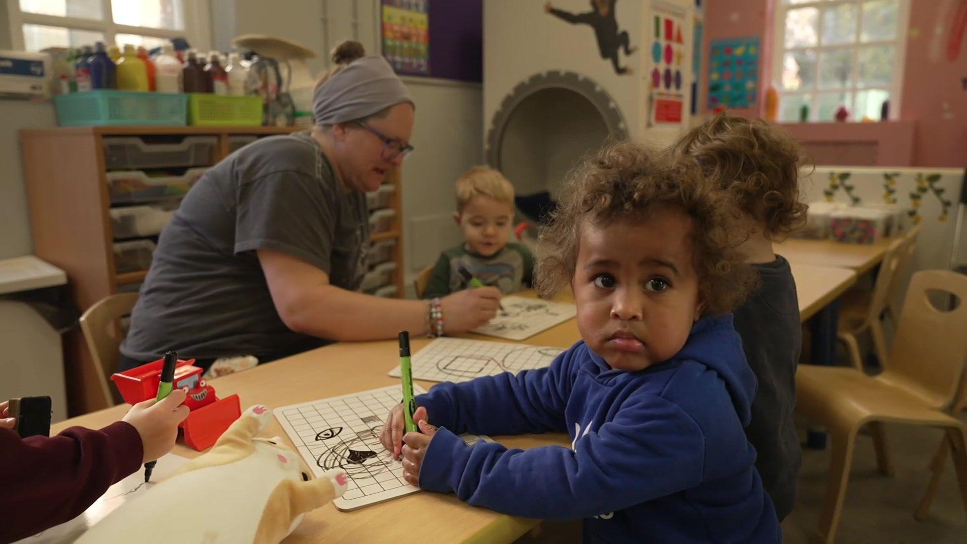 A group of children are sitting at a table drawing on paper. A childminder is helping them.
