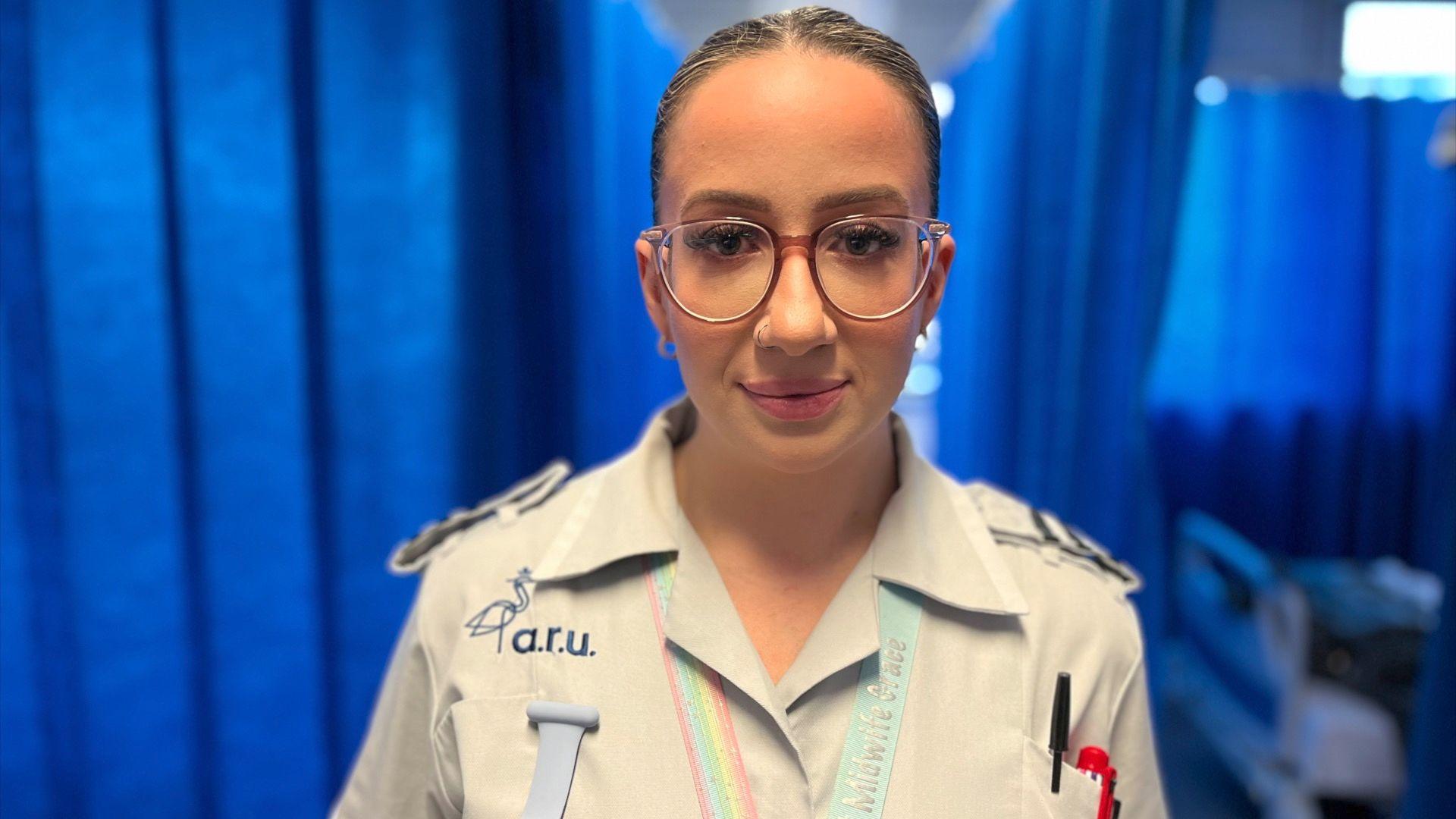 Grace Rungay, a trainee midwife at Basildon Hospital maternity unit, in her trainee scrubs, with a blue curtain on a ward behind her