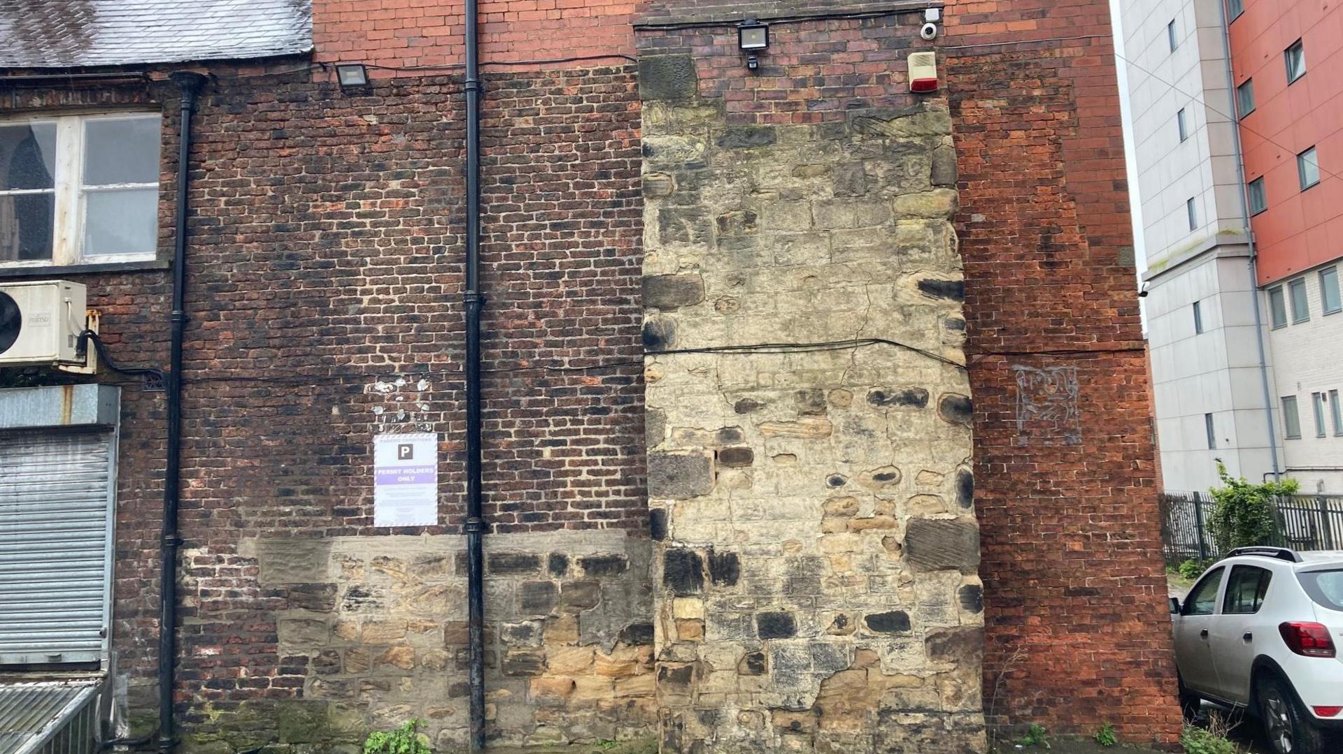 The wall of a property showing old brickwork with a white car parked nearby