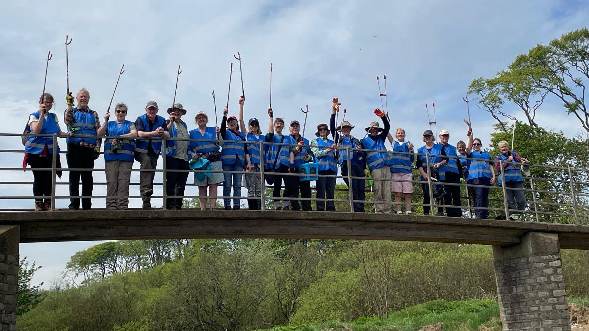 A row of people all holding litter pickers standing on a bridge 