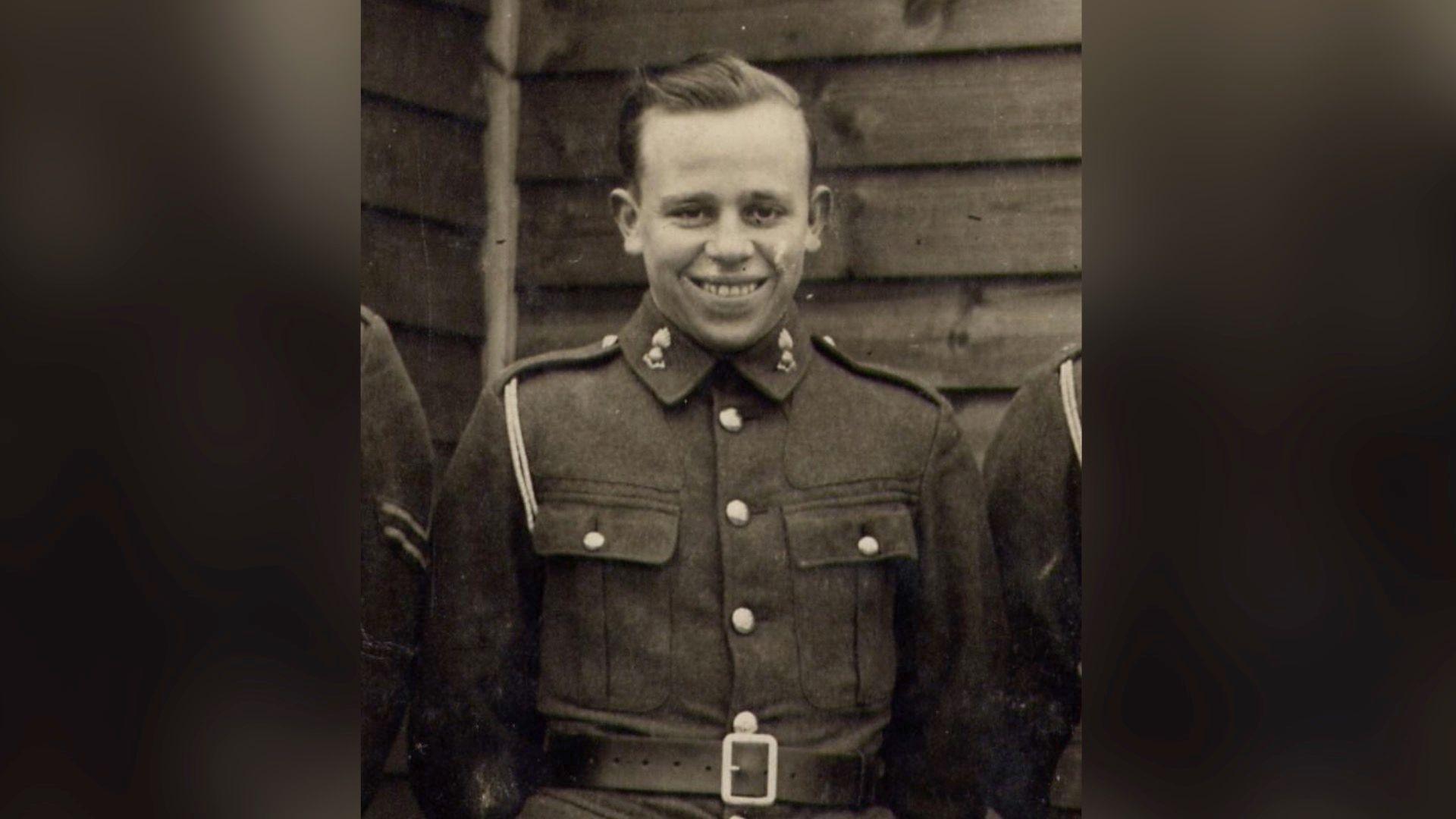 A man in an Army uniform. There are shoulders of two people either side of him. He is smiling and has brown hair.