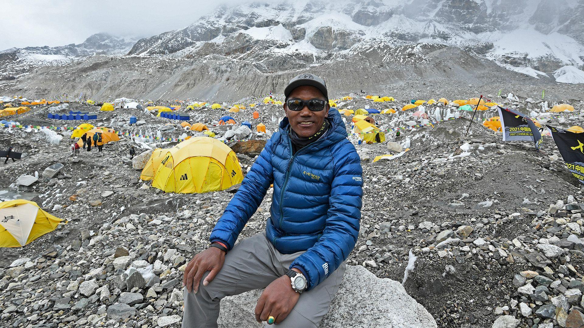 Kami Rita sits on a rock at the Everest base camp in the Mount Everest region of Solukhumbu district. There are lots of tents behind him and you can see the mountains in the background