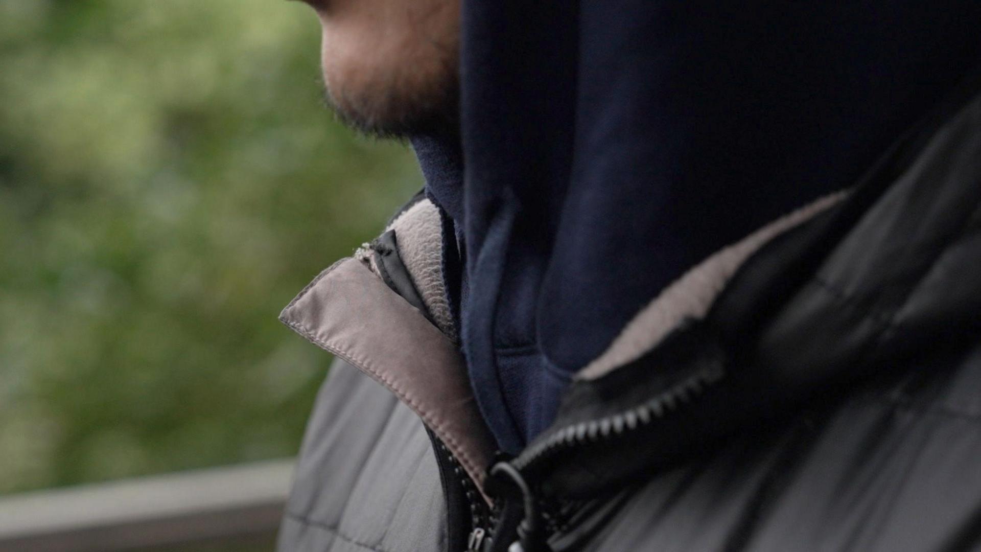 A person wearing a dark jacket shown against a soft-focus outdoor background. He appears to be photographed from chest up, shown in natural lighting with an urban setting behind him.