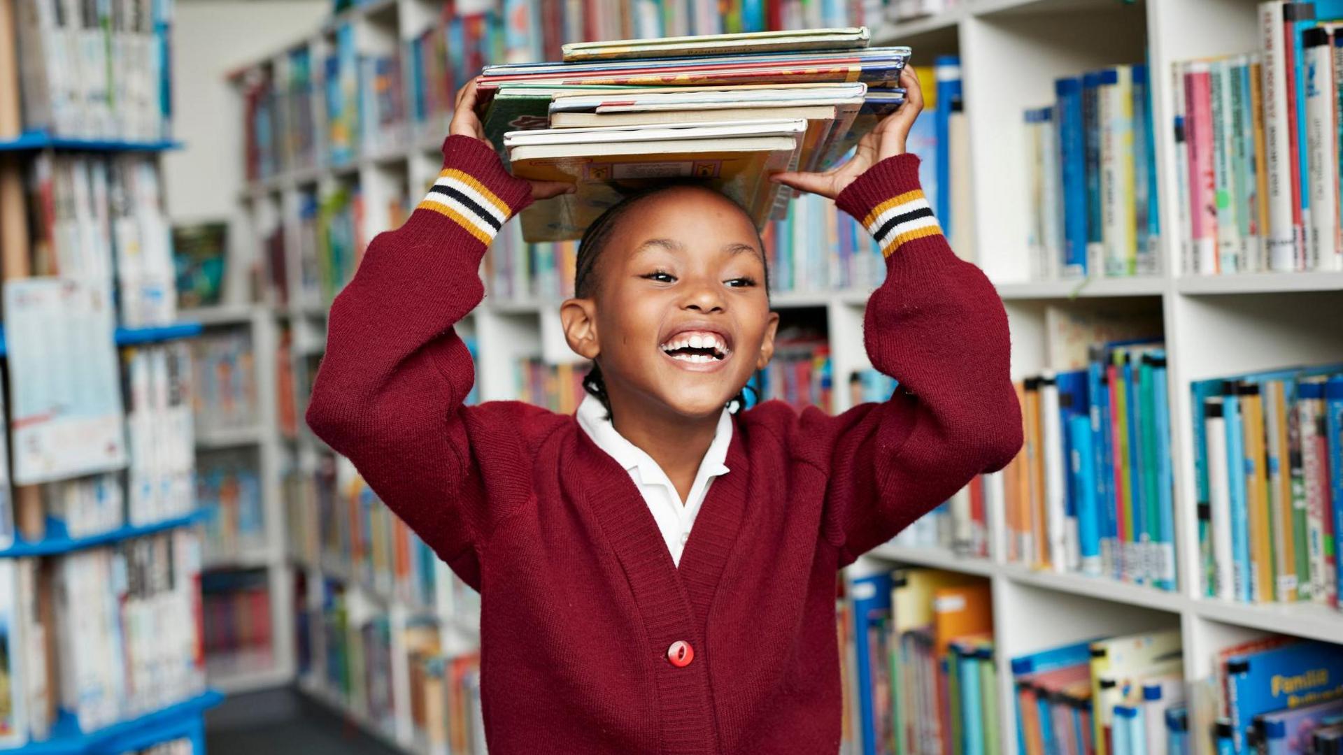 girl with a pile of books on her head in a library smiling and laughing