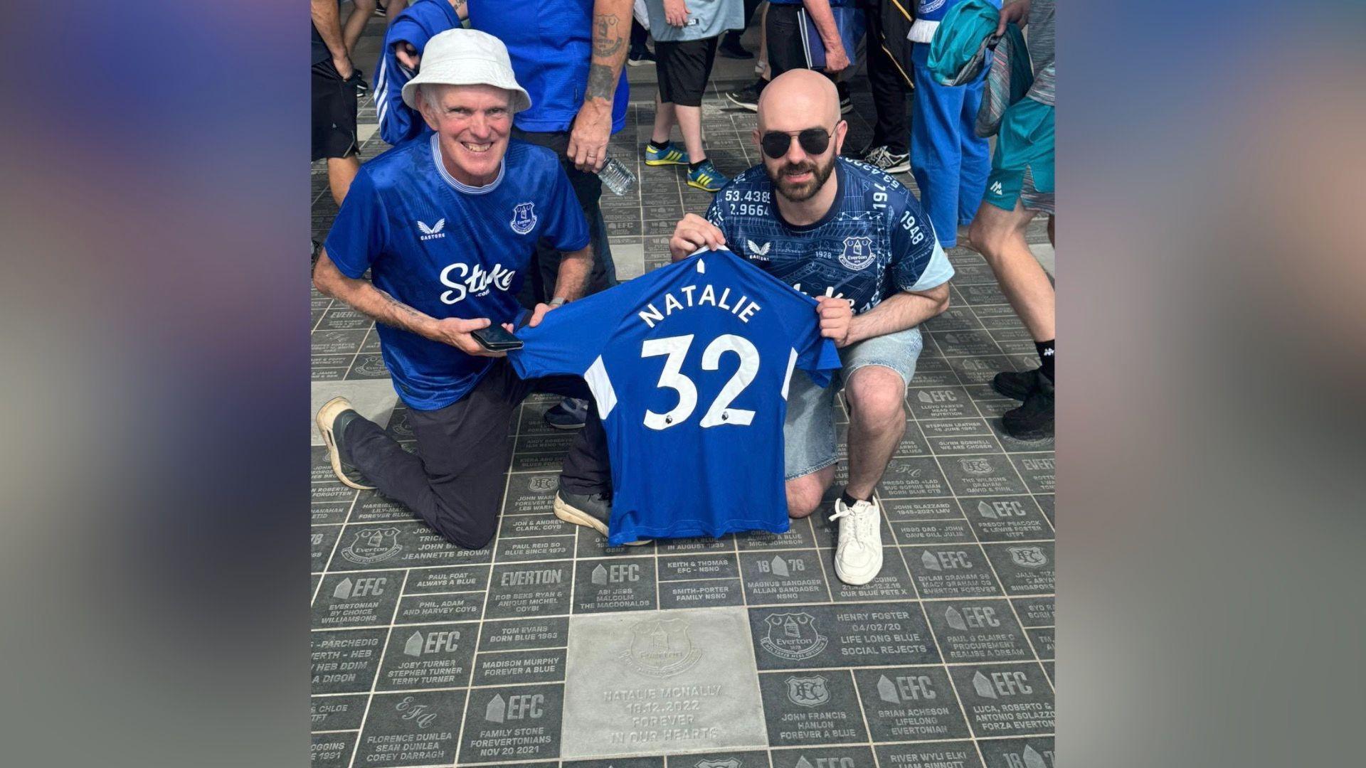 Noel and Declan are crouched down and holding a blue Everton jersey that says Natalie. One of the men is wearing a hat and the other is wearing sunglasses. A crowd is behind them.