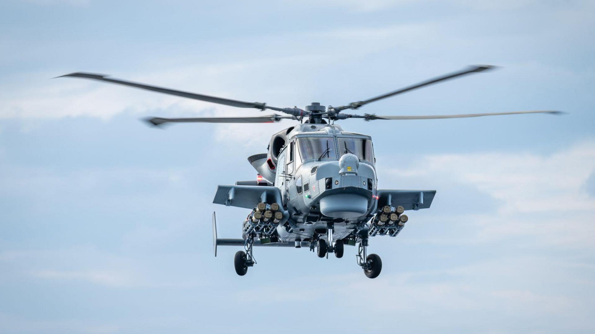 A grey Royal Navy Wildcat helicopter in flight, equipped with the Martlet missile system. The picture is a close up and the helicopter's rotor's are spinning.