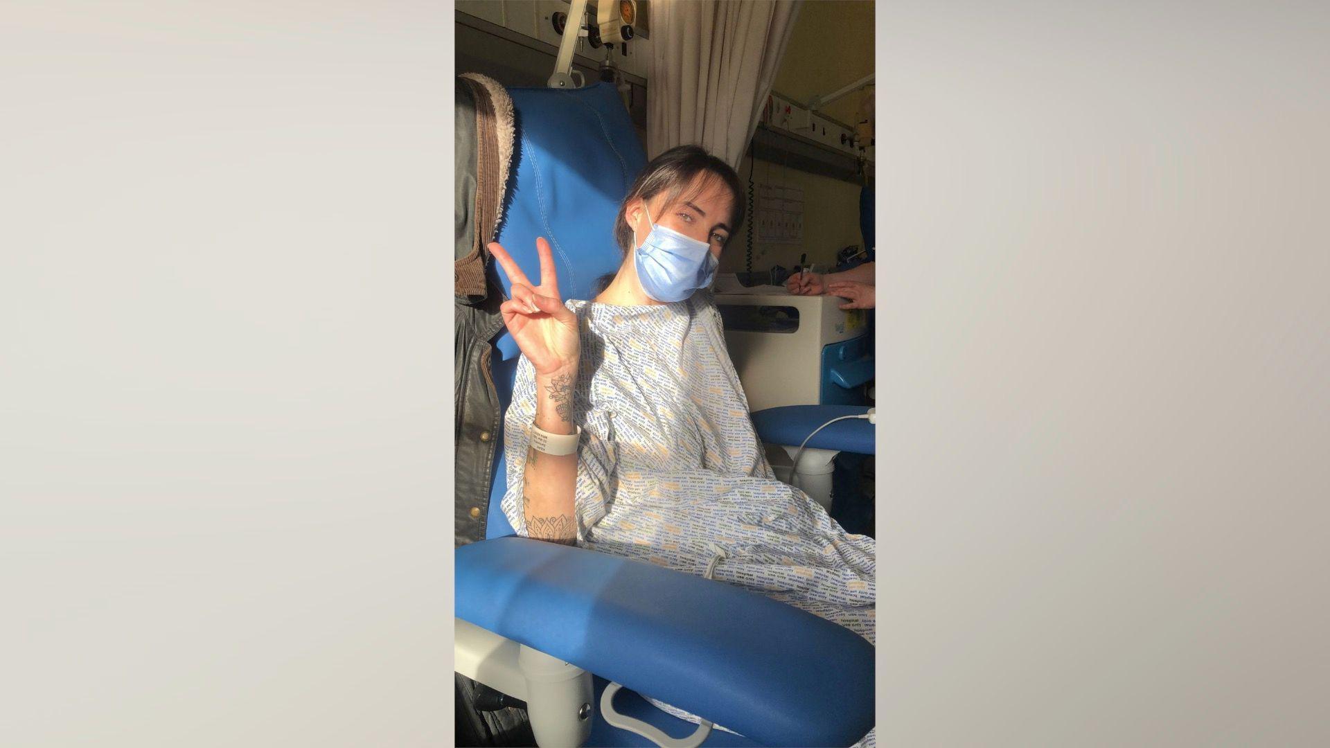 Sammy gives the peace sign with her hand as she sits in a hospital bed wearing patient clothes. She has a face mask on too and her brown hair is tied into a ponytail and the bed is in an upright position with blue cushions.