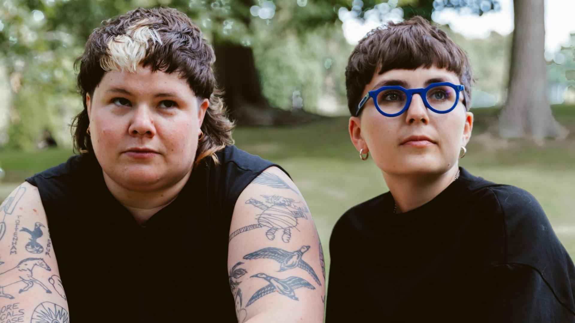 ARXX, made up of (L-R) Hanni Pidduck and Clara Townsend. Hanni has short brown hair with a blonde streak in their fringe and has heavily tattooed arms. Clara also has short brown hair with a fringe and wears cobalt blue rimmed round glasses. They're both dressed in black and are pictured sitting outside in a woodland area.
