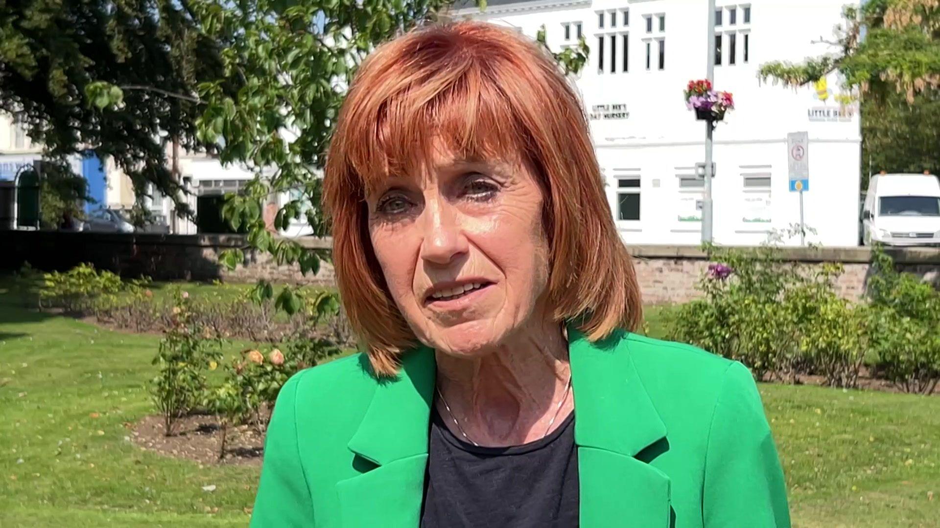 Woman with red hair, black top and green jacket standing in front of a park and a white building.