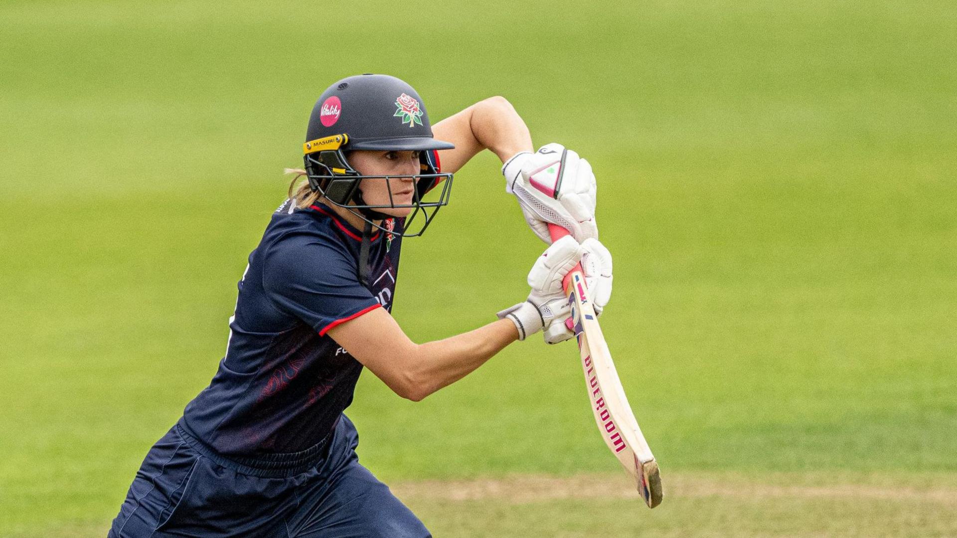 Gaby Lewis playing a shot off the front foot with her bat held high. She is wearing a navy kit with a navy helmet