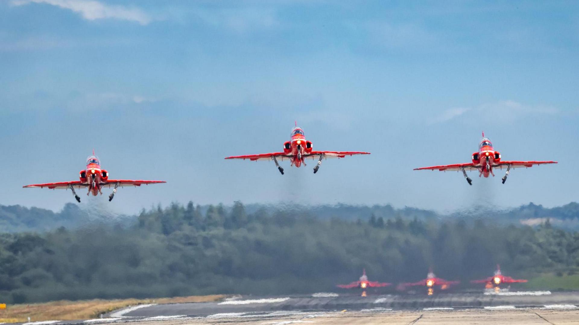 Three red jet planes take-off simultaneously with vapour trails behind them. Through those trails the image of the next three planes waiting can be seen on the runway behind them.