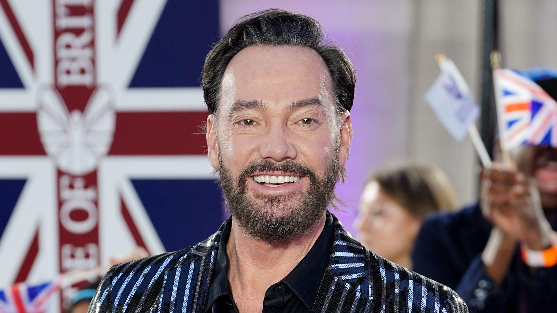 Craig Revel Horwood with black swept-back hair smiles at the camera at the Pride of Britain Awards. He has a beard and a sequined black jacket.