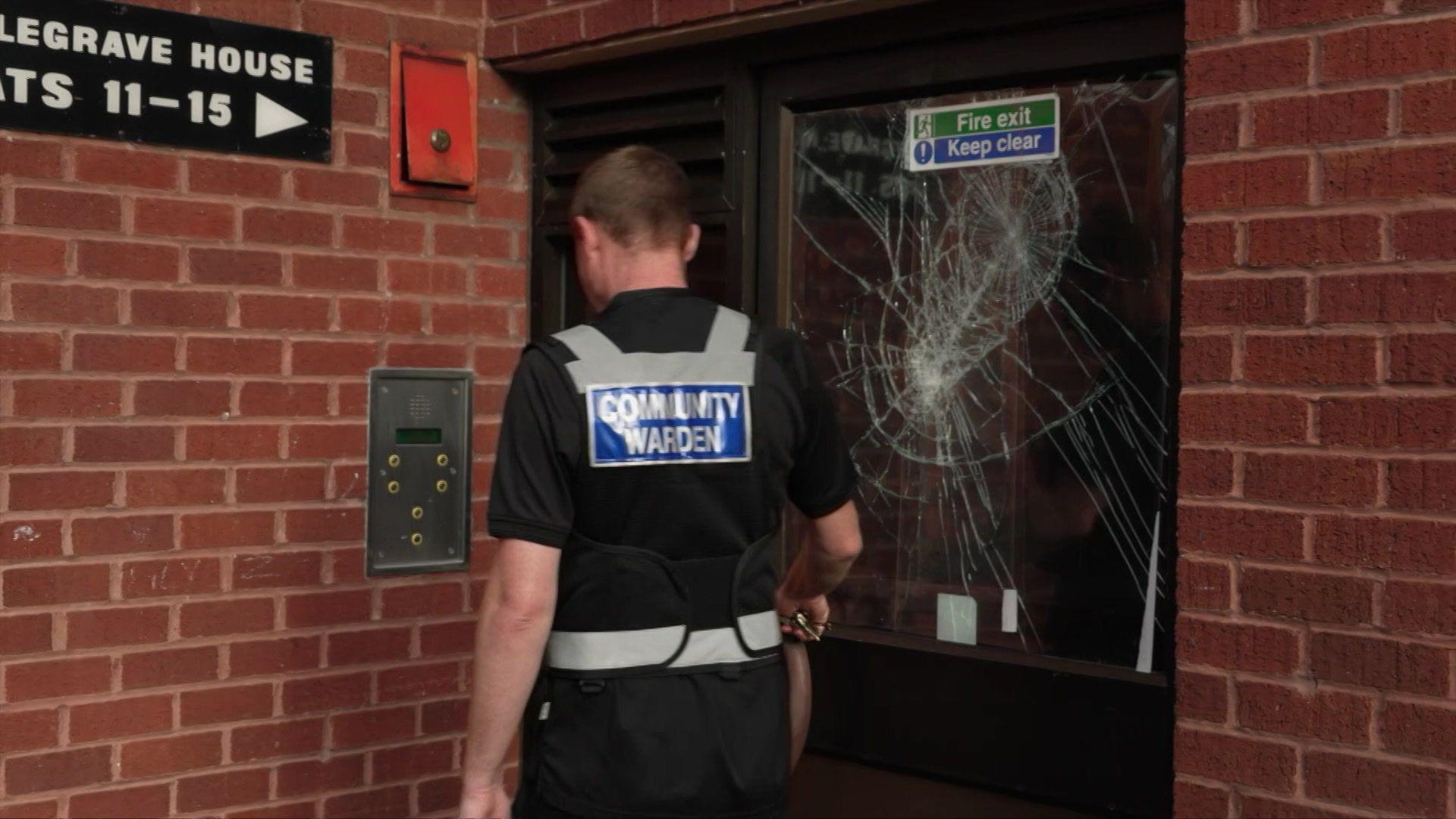 The back of a community warden who is walking towards a door with a smashed window.