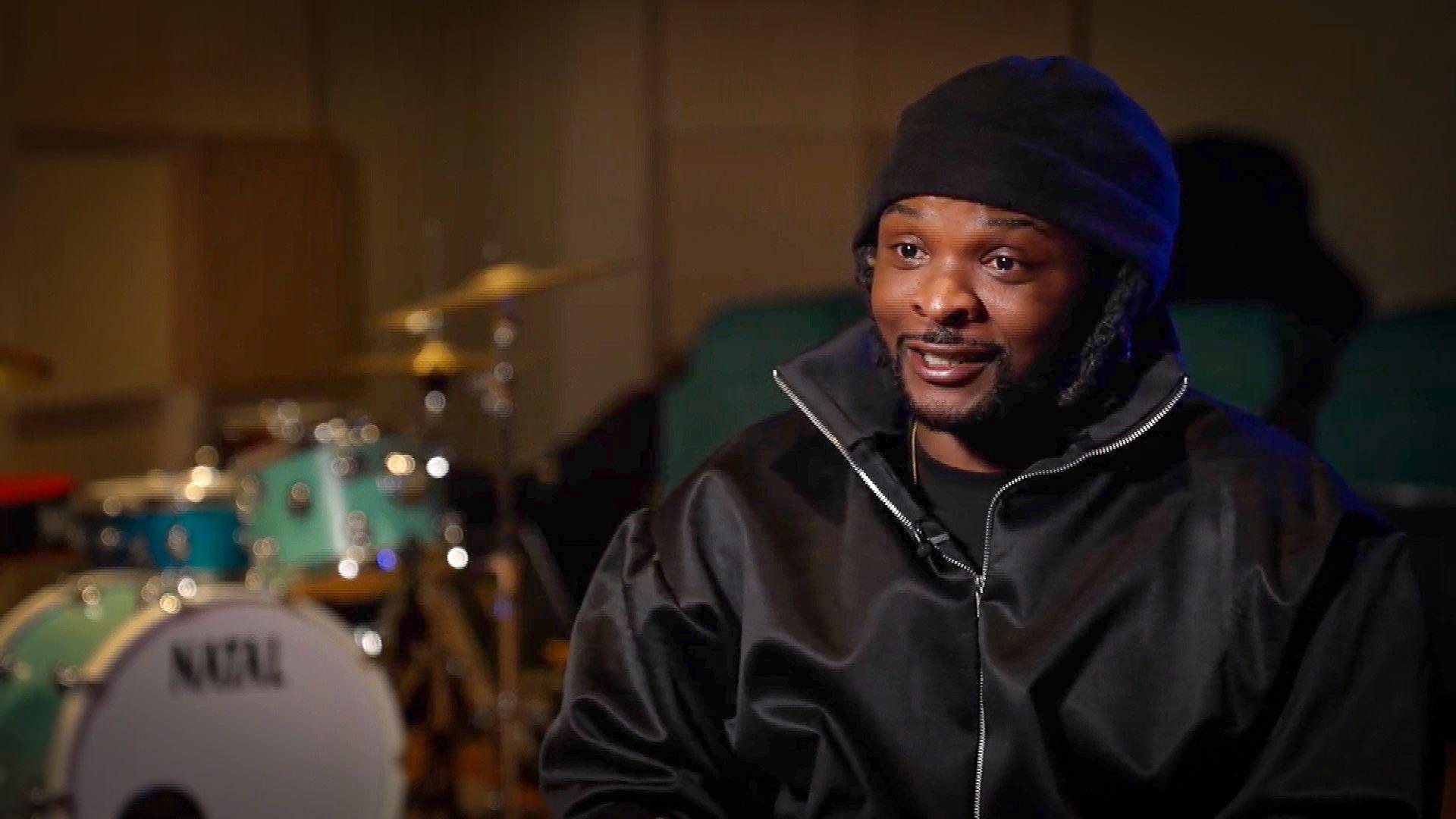 Rosca Onya in a music studio wearing black. A drum kit can be seen behind him, out of focus.