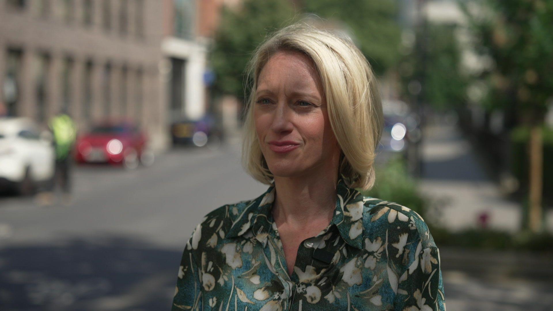Claire Waxman, with shoulder-length blonde hair and wearing a dark green shirt with white flowers on it is standing on a London street