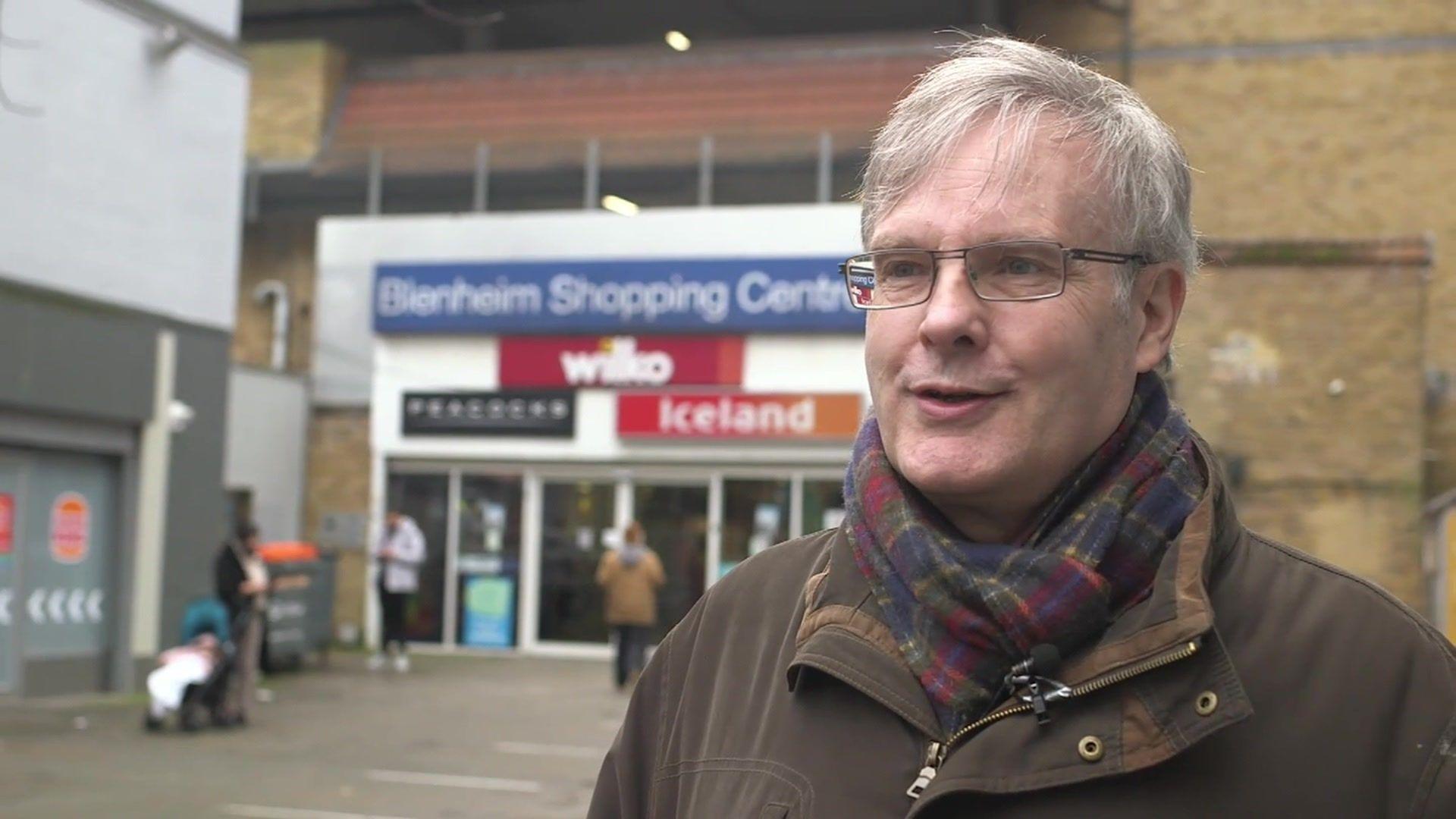 Mike Payne, a man with grey hair, a brown coat and checked scarf, standing in front of the shopping centre