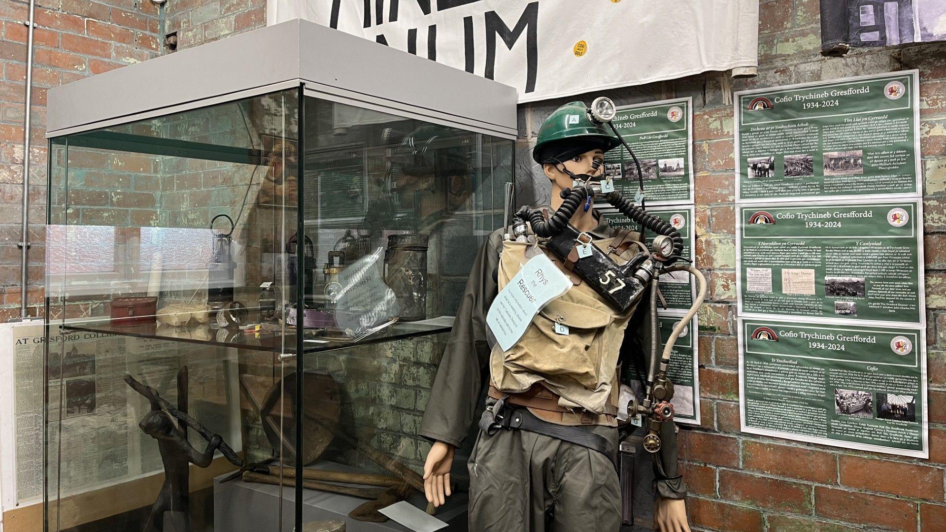 A mannequin wearing the kind of kit which would have been worn by the mine rescuers, khaki clothing and a dark green hard hat and breathing apparatus. There is a display cabinet to its left with some mining memorabilia and to its right, a brick wall with information boards about pits in north Wales. 