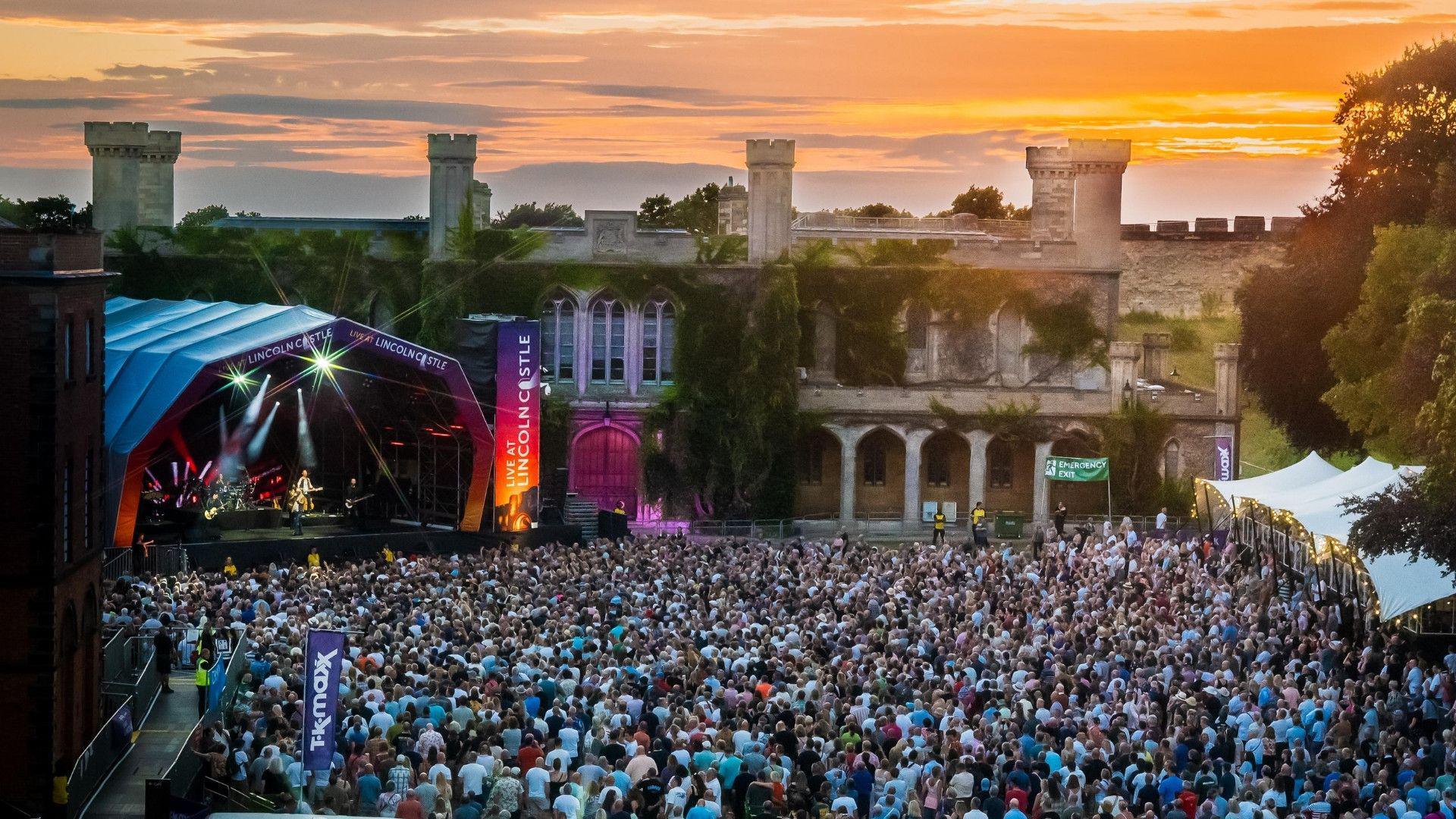 A large crowd of people are gathered in front of a large stage next to a Gothic building with greenery on its walls. Someone can be seen performing on the stage with the words 'Live at Lincoln Castle' written around it.
