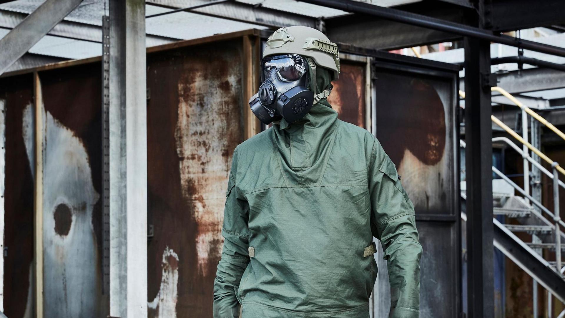 A figure wearing a large gas mask with green helmet and green overall with industrial background.