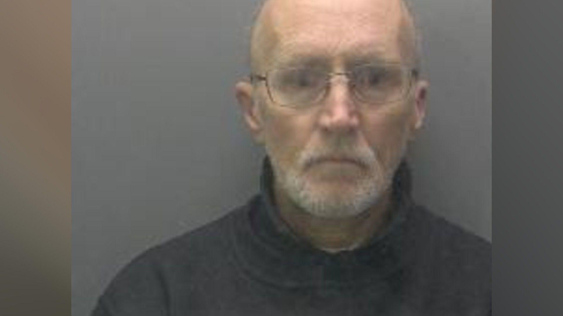 Peter Swan's custody photo. He is wearing a black or navy turtle neck jumper. He is bald and has glasses on.