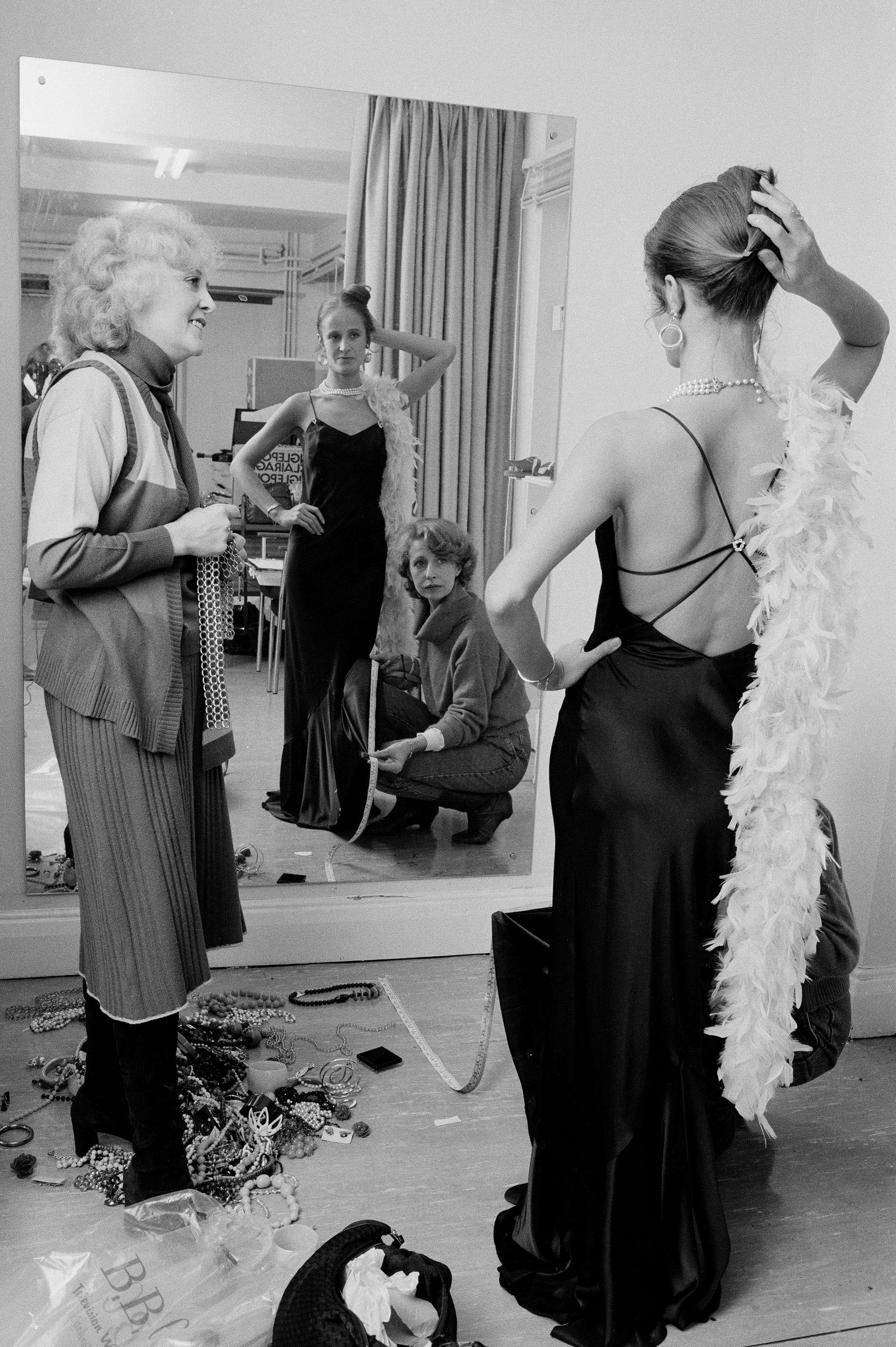 jean fennell at a costume fitting, wearing a black backless dress and feather boa  looking in the mirror