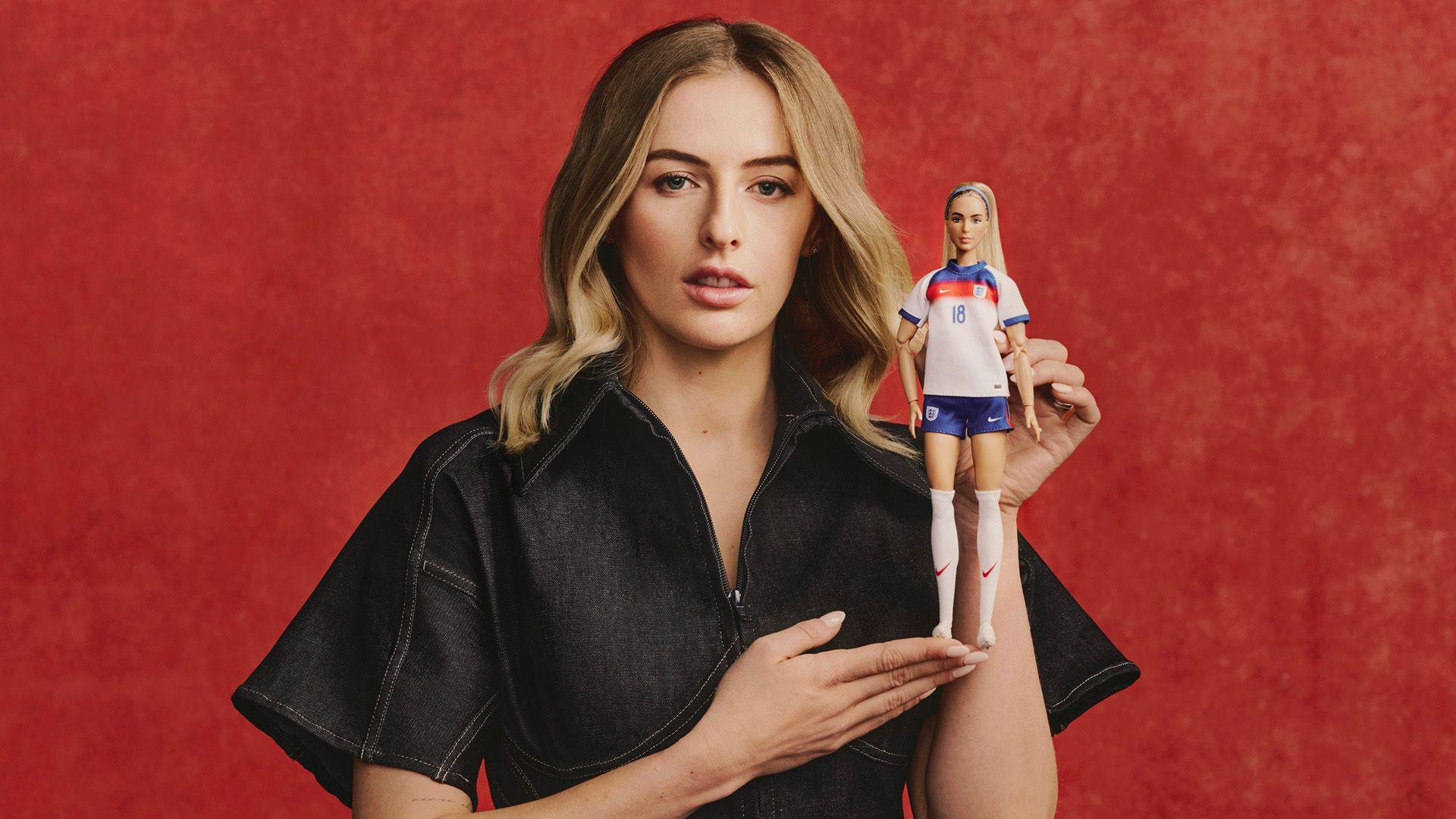 Chloe Kelly wears a black shortsleeve jumpsuit. She is holding up her Barbie doll. Her hair is short, blonde and in loose waves. 