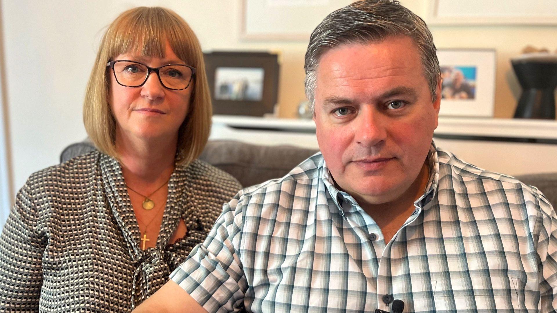 A picture of Melanie and Stephen Cull, both wearing black and white patterened shirts, sitting on a grey sofa with pictureframes in the background