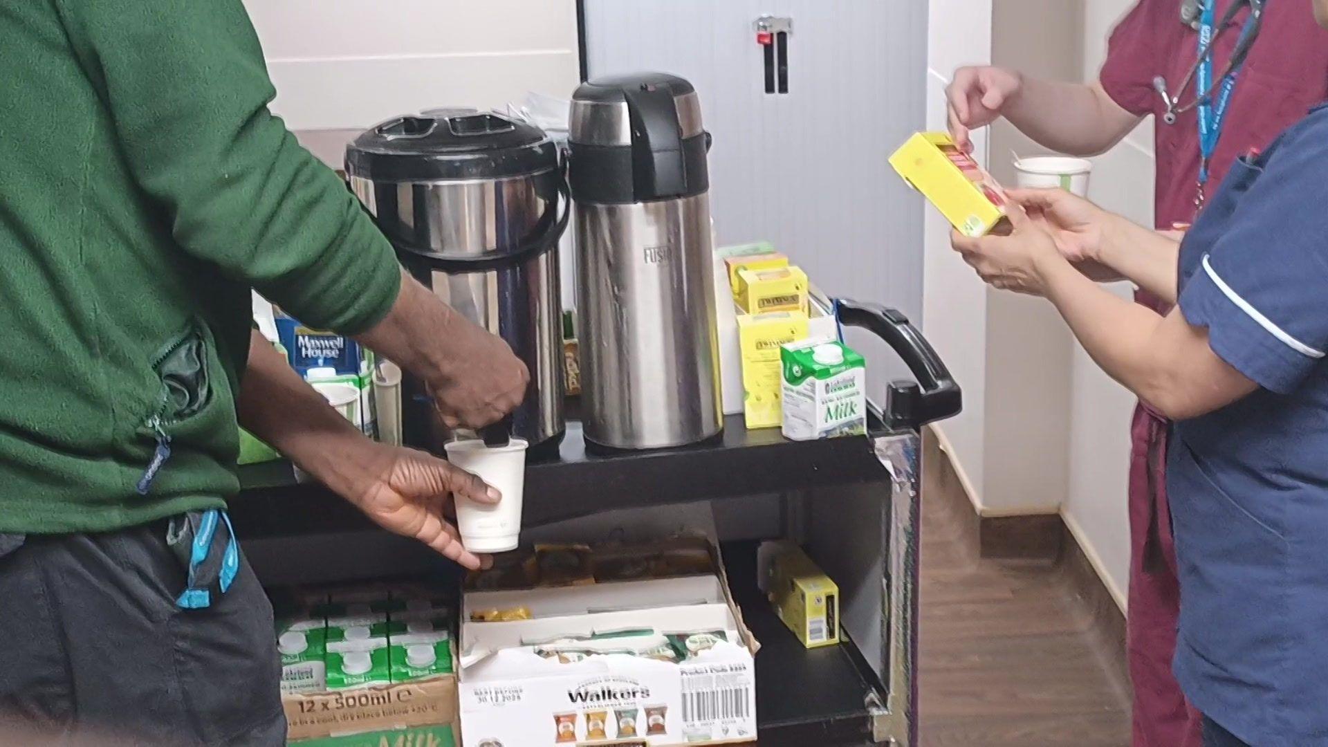 Tea trolley in action at the hospital - a man is pouring hot water from the large mettle into a white cup. He is wearing a dark green sweater and navy trousers. A woman in nurse's uniform is looking at a box of tea.