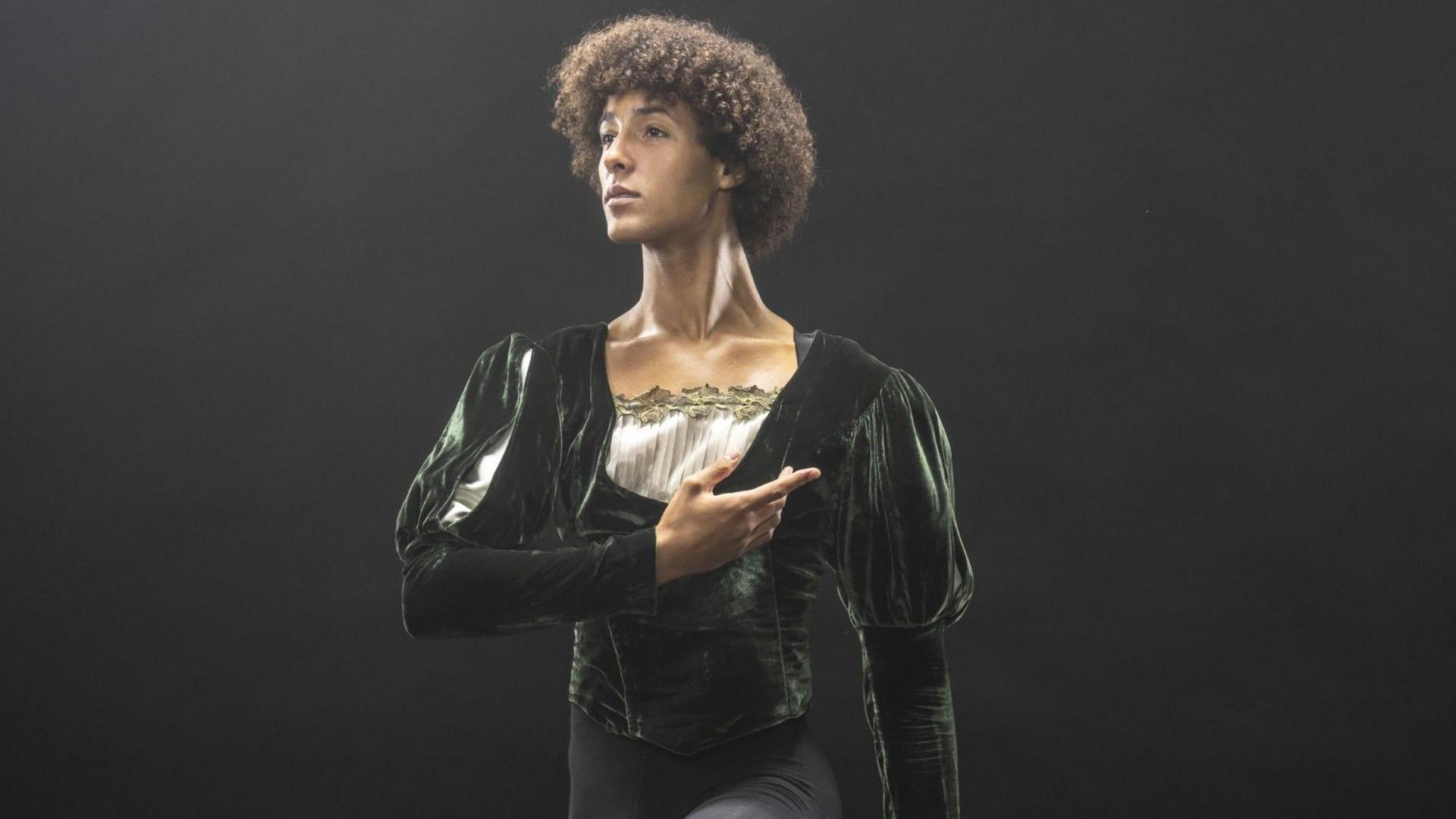 Jakob, in a ballet poise, with one arm across his body, wearing a dark ballet outfit, with velvet top and black tights, in front of a black wall. He has short dark curly hair. 