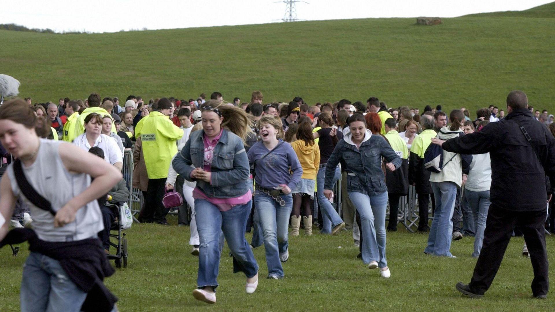 Several young women run into a field after being let through a security gate behind which a crowd is gathered. A man in black is gesturing at them to slow down.
