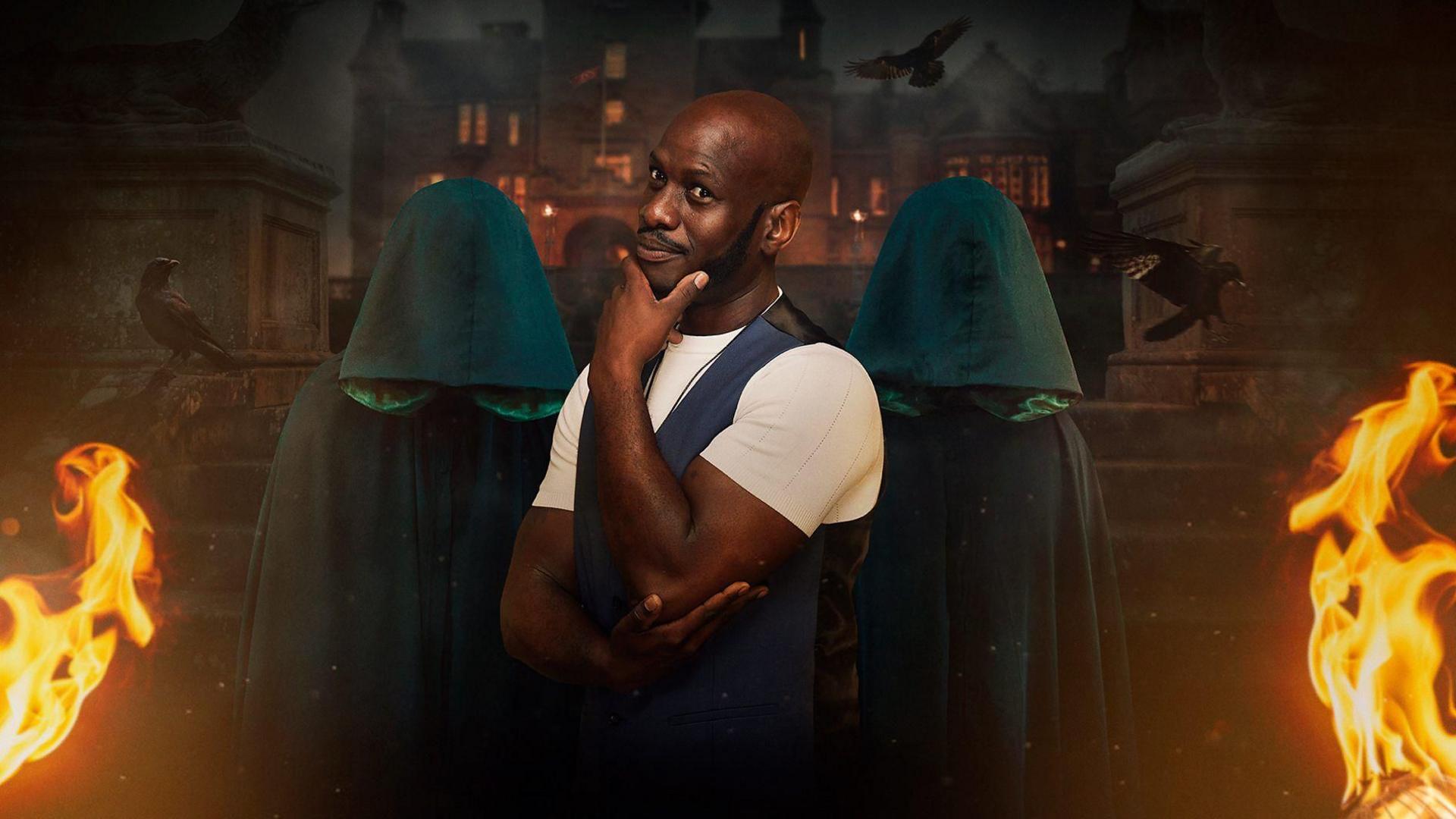 A man stands in front of a backdrop showing a castle, cloaked figures, crows and flames. He is wearing a white t-shirt and a blue waistcoat. He has a shaved head and a black goatee.