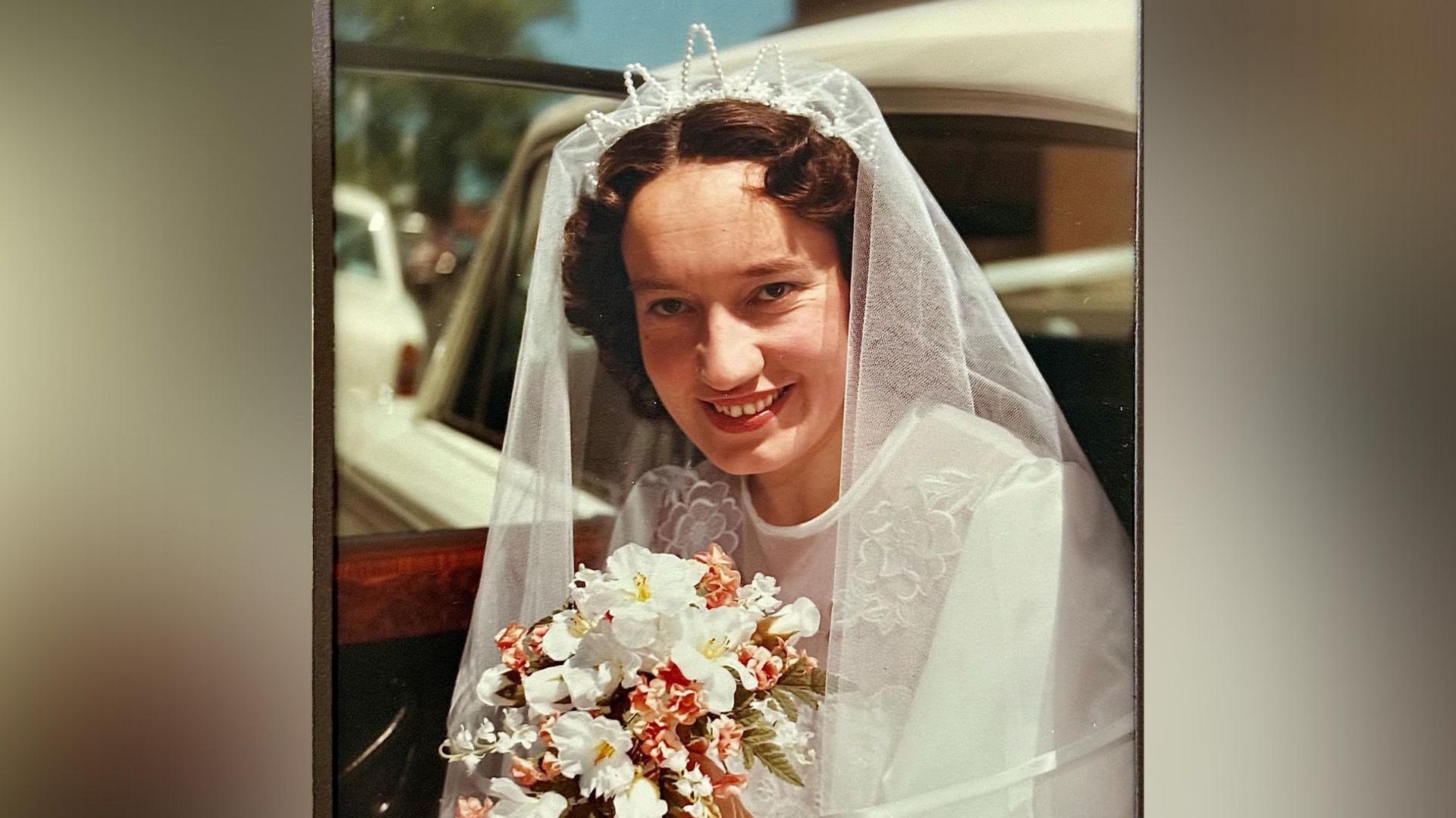 A photograph of a bride with curly brown hair pinned up smiling towards the camera. She wears a typical white dress and veil and holds a white and pink bouquet. She is pictured in the doorway of a white car.