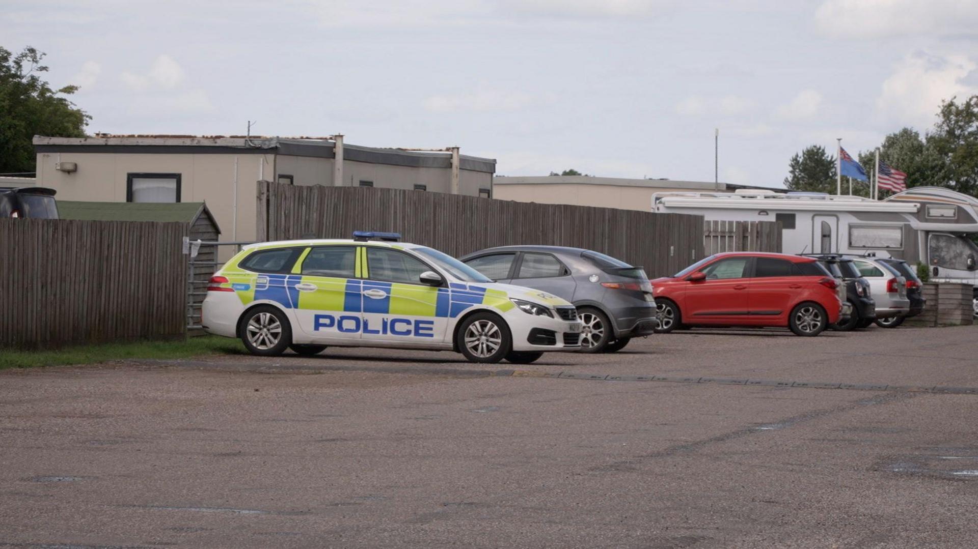 Police car parked in a car park. Other vehicles are also parked in the car park. A wooden fence runs around the carpark.