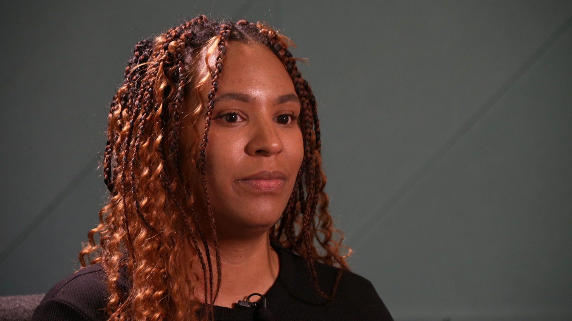 Lauren Caulfield, who has brown, braided hear. She wears a neutral expression as she talks to a journalist.