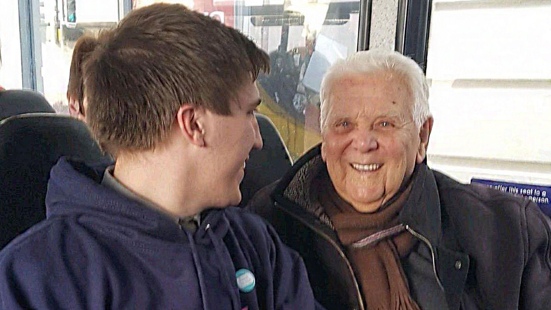 A young man in a hoodie talking to an older man in a black coat and brown scarf on a bus.