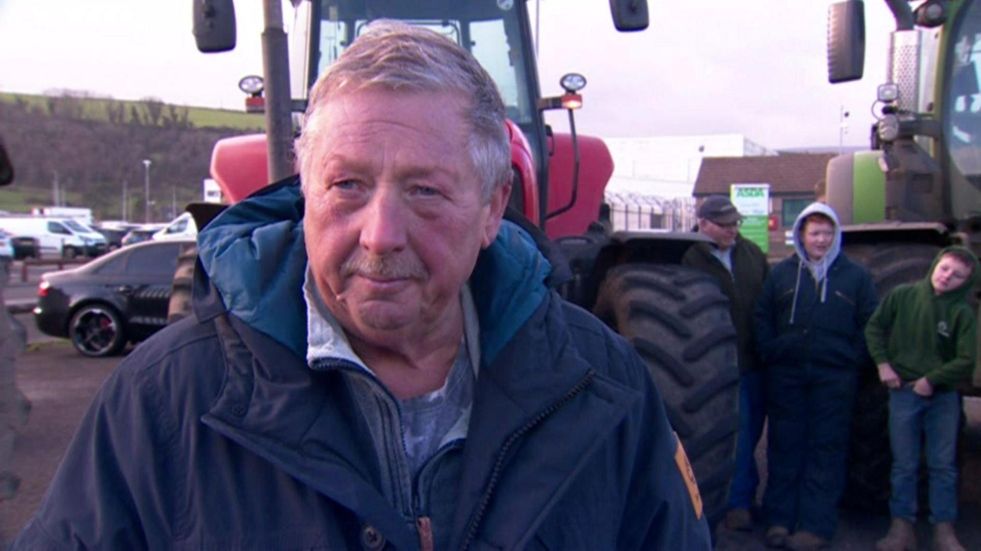 Sammy Wilson stands in front of a red tractor. He has grey hair and a blue coat. An Asda sign is visible behind him.