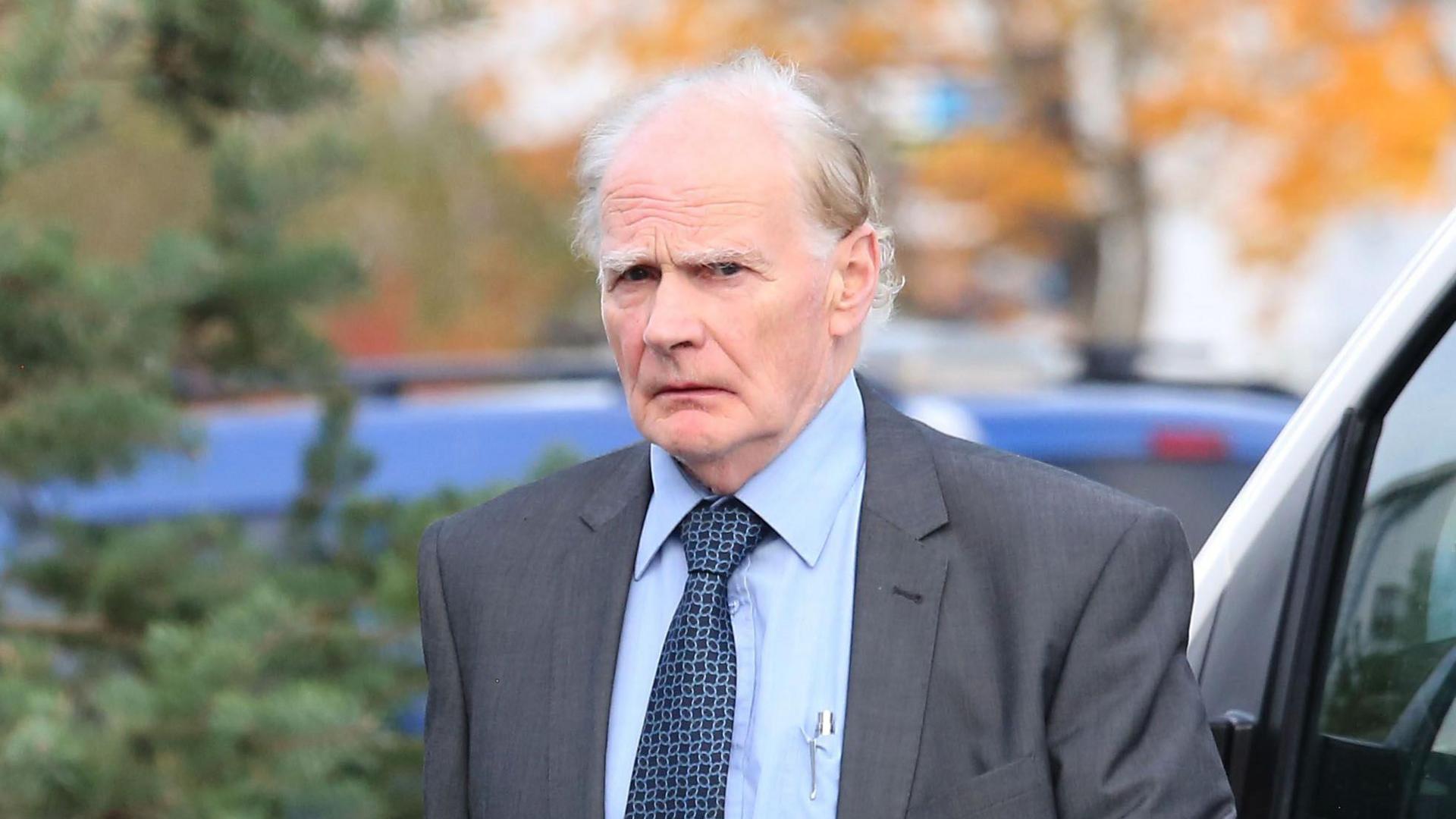 A photo of Douglas Stewart taken outdoors. He looks towards the camera at a slight angle. He is wearing a grey suit, blue shirt and tie. He is bald on top with grey hair at the side and thick grey eyebrows.